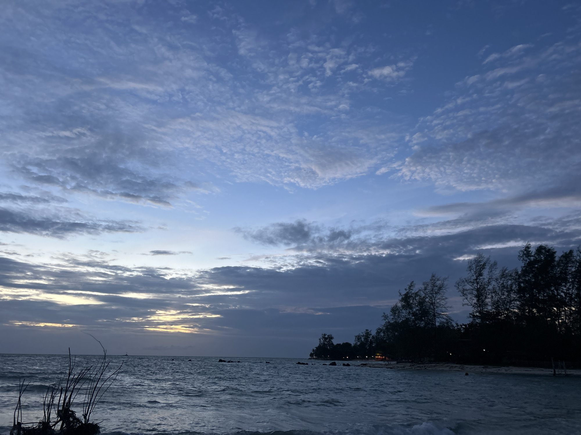 This is a photograph of a beach scene at dusk in Ko Pha-ngan District, Thailand. The sky is a deep blue with scattered clouds. The sun appears to be setting or has just set, with a sliver of yellow light visible on the horizon. The ocean is a dark blue with moderate waves. In the foreground, on the left side of the image, there is a cluster of dark, dried branches and debris. On the right side, silhouetted trees line the beach, with some lights visible amongst them, suggesting human habitation or activity. The overall atmosphere is serene and tranquil, typical of a sunset on a tropical beach.