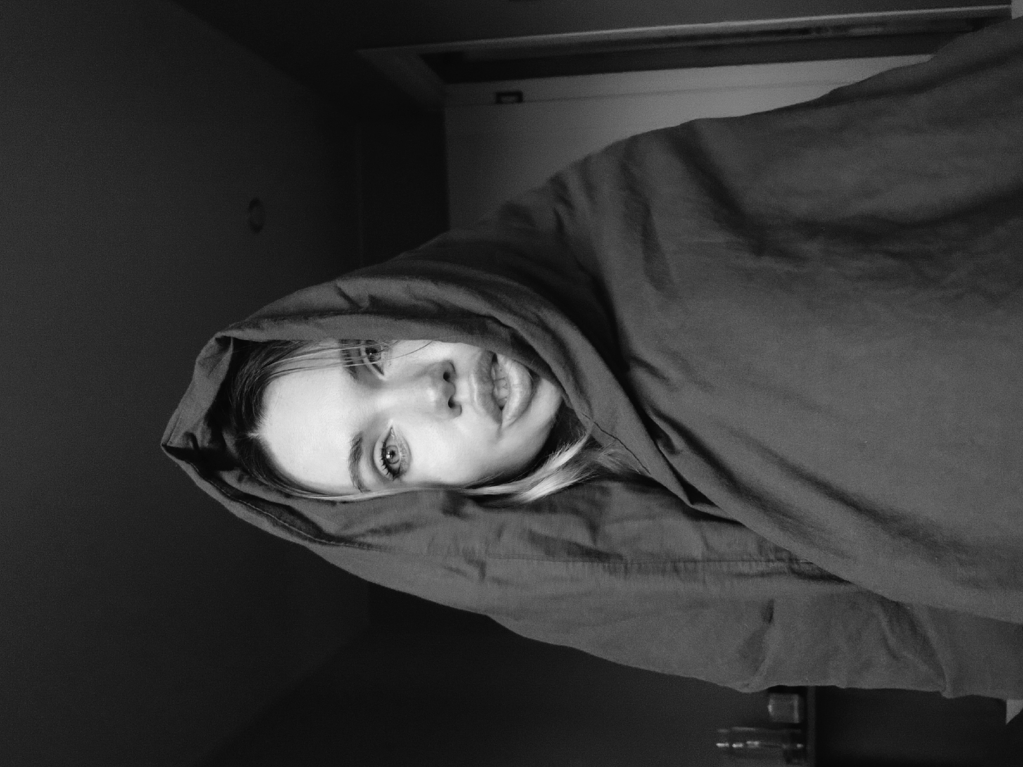 A black and white portrait shows a fair-skinned person with light eyes and light hair partially covered by a dark fabric, possibly a blanket or shawl, which forms a hood around their head. The person is looking directly at the camera. The setting appears to be indoors, with a dark wall in the background on the left and a door or other architectural feature visible at the top. The lighting is dim, creating shadows and highlighting the subject's face. The image is taken from a slightly low angle, with the subject positioned off-center. The photograph does not depict a location in Votkinsk, Russia, but rather is an indoor shot of a person in a domestic setting.