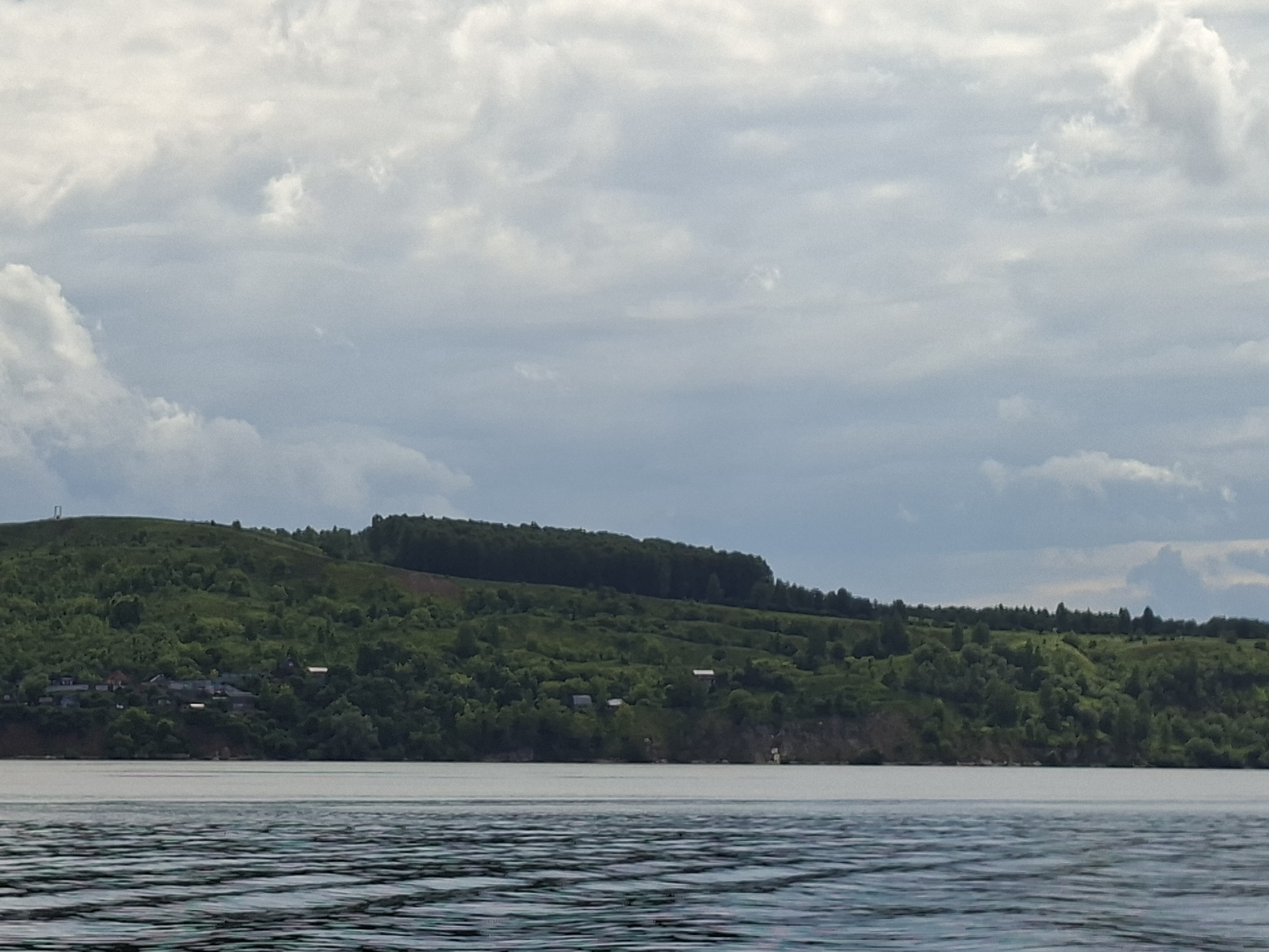 The image depicts a panoramic view of a body of water in the foreground, with a lush green, rolling hillside in the background. The hillside is densely covered in trees, with a few scattered houses and buildings visible along its base. The sky above is filled with dramatic, layered clouds, suggesting a dynamic weather system. The overall scene evokes a sense of natural beauty and tranquility, likely capturing a moment on the Volga River near Kazan, Russia, a region known for its scenic landscapes.