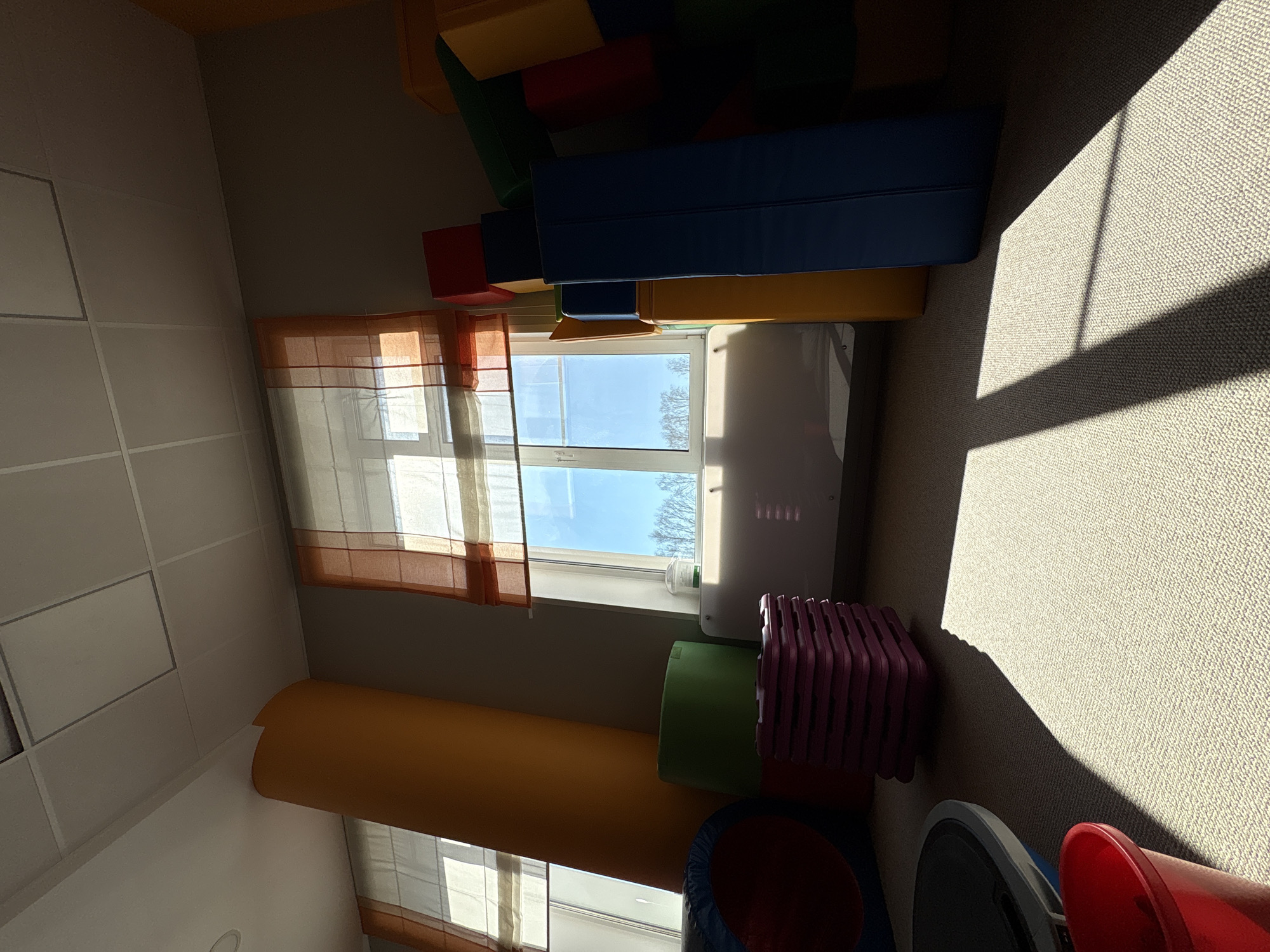 The image shows an indoor playroom in Izhevsk, Russia, featuring a window with orange-trimmed blinds, a stack of colorful foam blocks and cylinders, a blue tunnel, pink stackable chairs, and a balance board. Strong sunlight creates shadows on the beige carpet. There are no people visible in the frame.