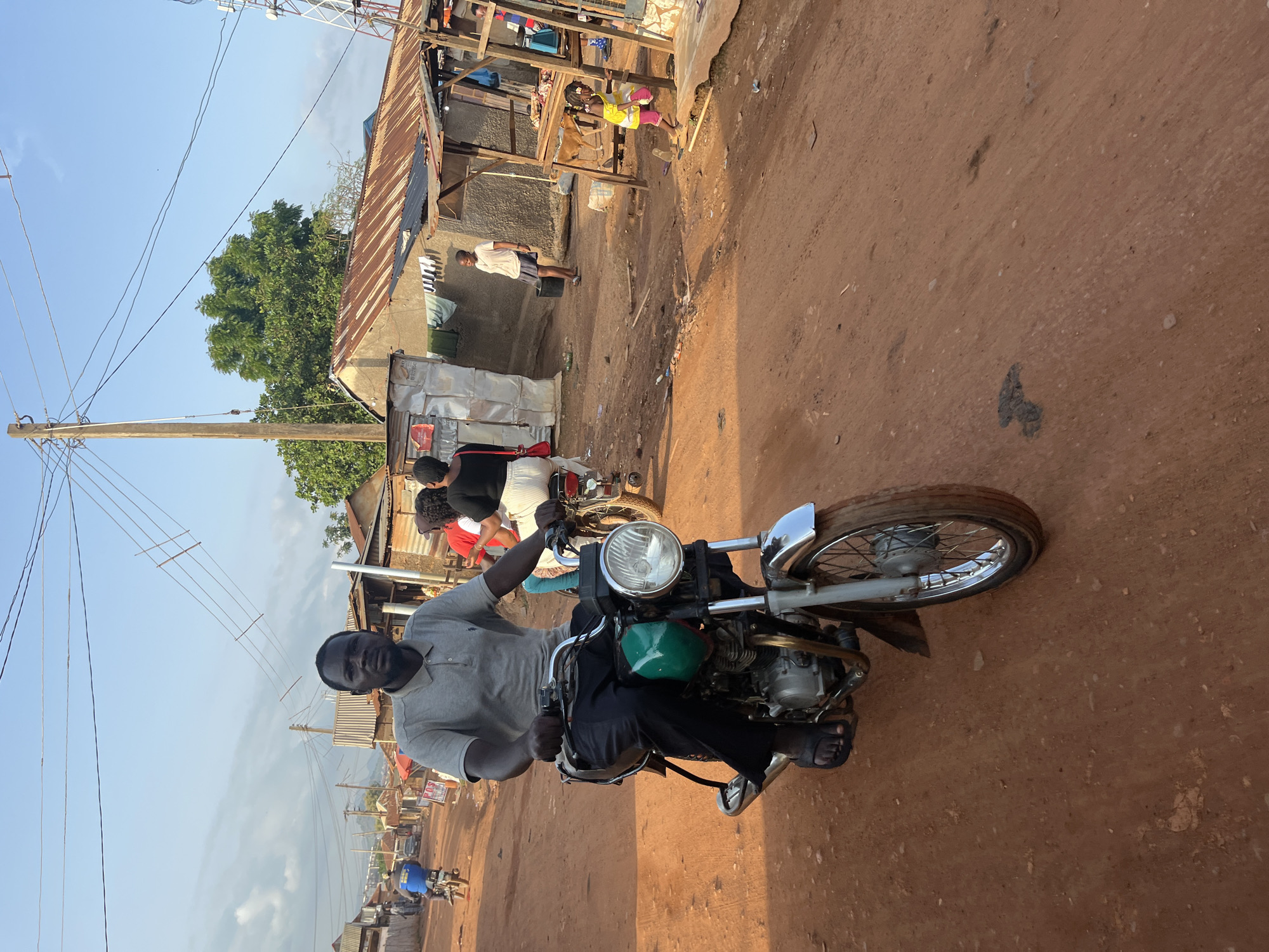 A man wearing a grey polo shirt and black trousers rides a motorcycle on a dirt road in Abuja, Nigeria. The motorcycle is green and silver with a silver headlight. Behind the man, other people and motorcycles are visible. The background shows a settlement with tin-roofed buildings and trees. The sky is blue with some clouds.