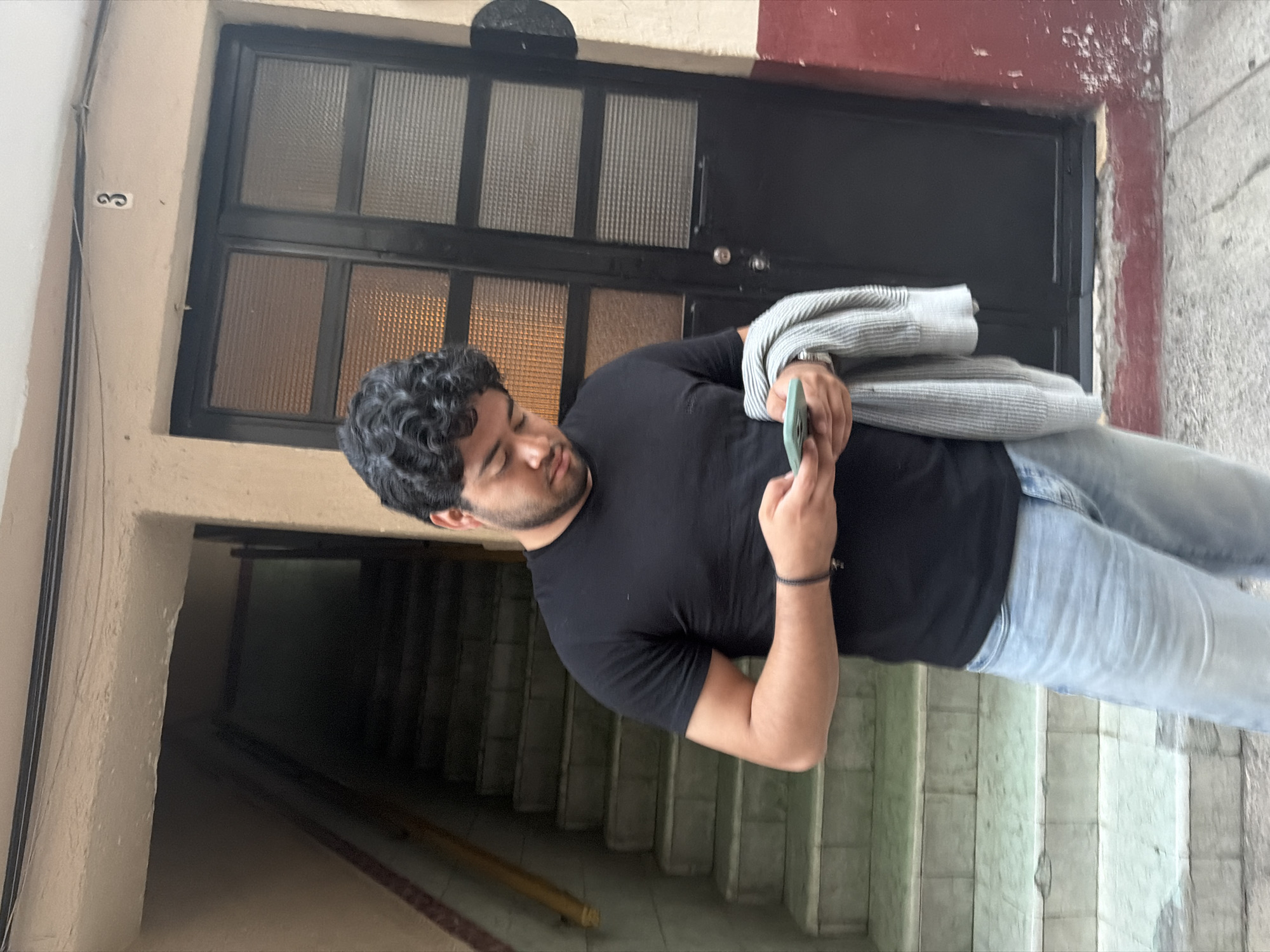 A medium shot, composed at an oblique angle, depicts a man using a cell phone while standing on the stairs of a building in Puebla, Mexico. The man has dark curly hair, a beard, and a dark complexion. He is dressed in a black t-shirt and light blue denim jeans, and wears a dark-colored wrist band on his right wrist. He is holding a cell phone in his hands, facing toward the left side of the frame. A grey and white striped garment is folded over his left arm. The background includes a dark-painted door with two rows of glass panes and a hallway leading to the interior. The stairwell features tiled steps. A number '3' is visible on the wall to the left. The top of the door frame is colored red. The lighting appears to be daylight.
