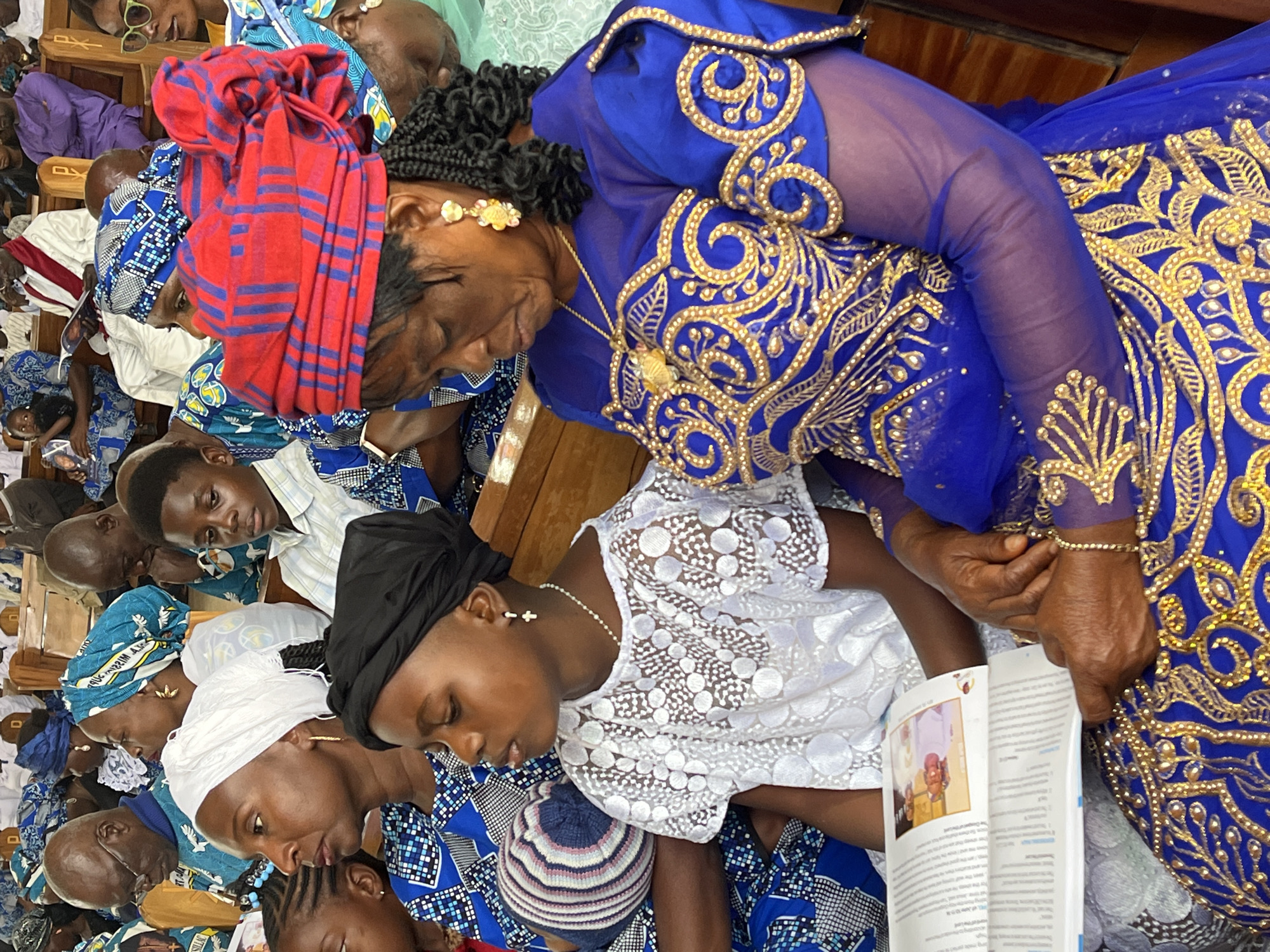 In a church setting, an elderly Black woman wearing a vibrant blue and gold embroidered dress and a red and blue striped headscarf is seated on a wooden pew. She is looking down at an open book held by her hands. Beside her, a young Black girl in a white lace dress and black headscarf is also seated, looking down. Other individuals, including men and women of various ages, are visible in the background, some wearing colorful headscarves and traditional African attire, suggesting a religious gathering. The overall atmosphere appears solemn and respectful. The location is identified as Vom, Nigeria.
