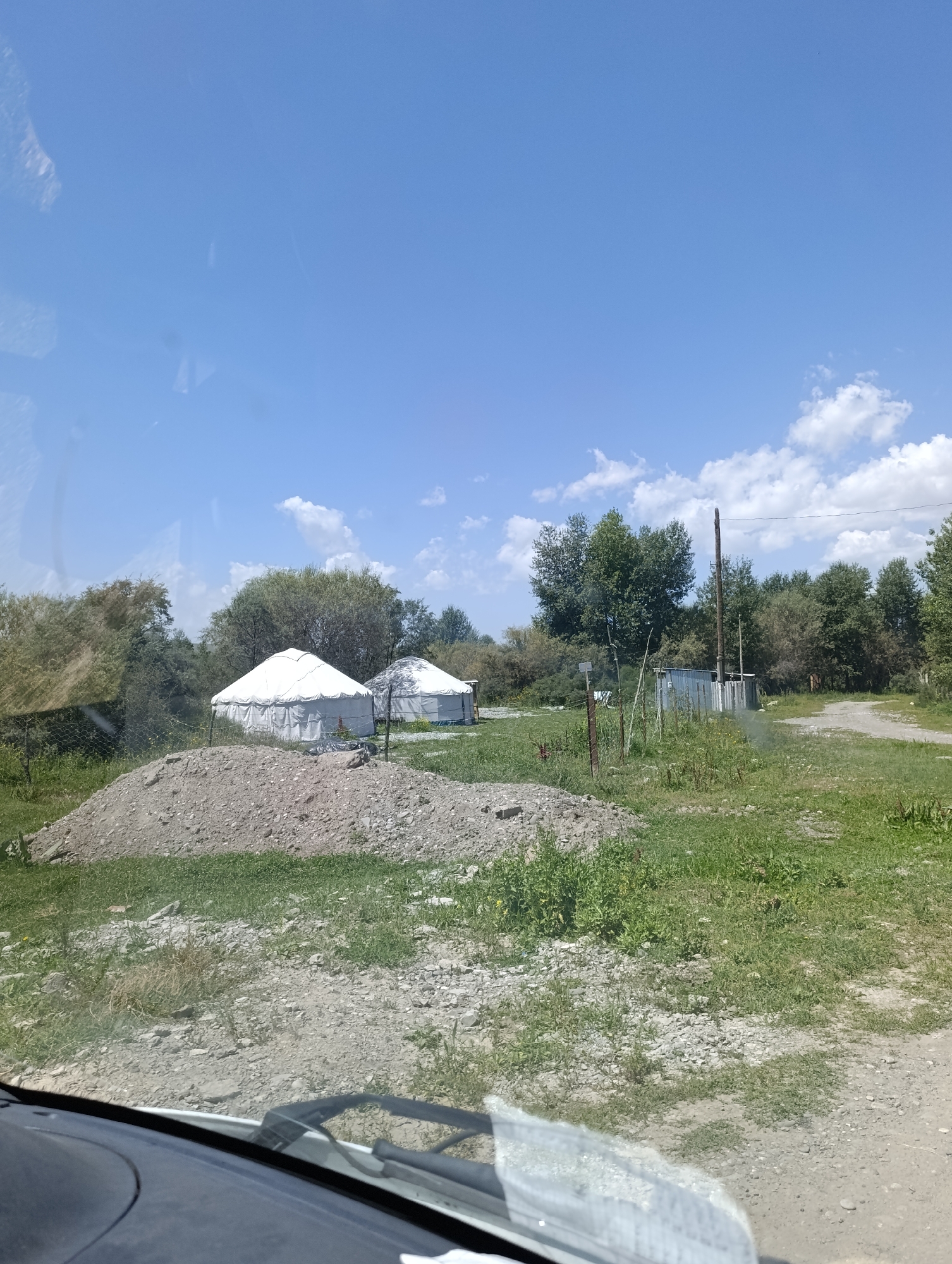 The image depicts an outdoor scene in the city of At-Bashy, Kyrgyzstan. In the foreground, a dirt road and a pile of rubble are visible. Beyond the rubble, there are two white yurt tents and some green vegetation. In the background, a line of trees forms a natural barrier. A metal structure, possibly a shed, with a wooden pole and wires, is situated to the right of the yurts. The sky is blue with scattered white clouds. The overall impression is that of a rural or semi-rural setting.