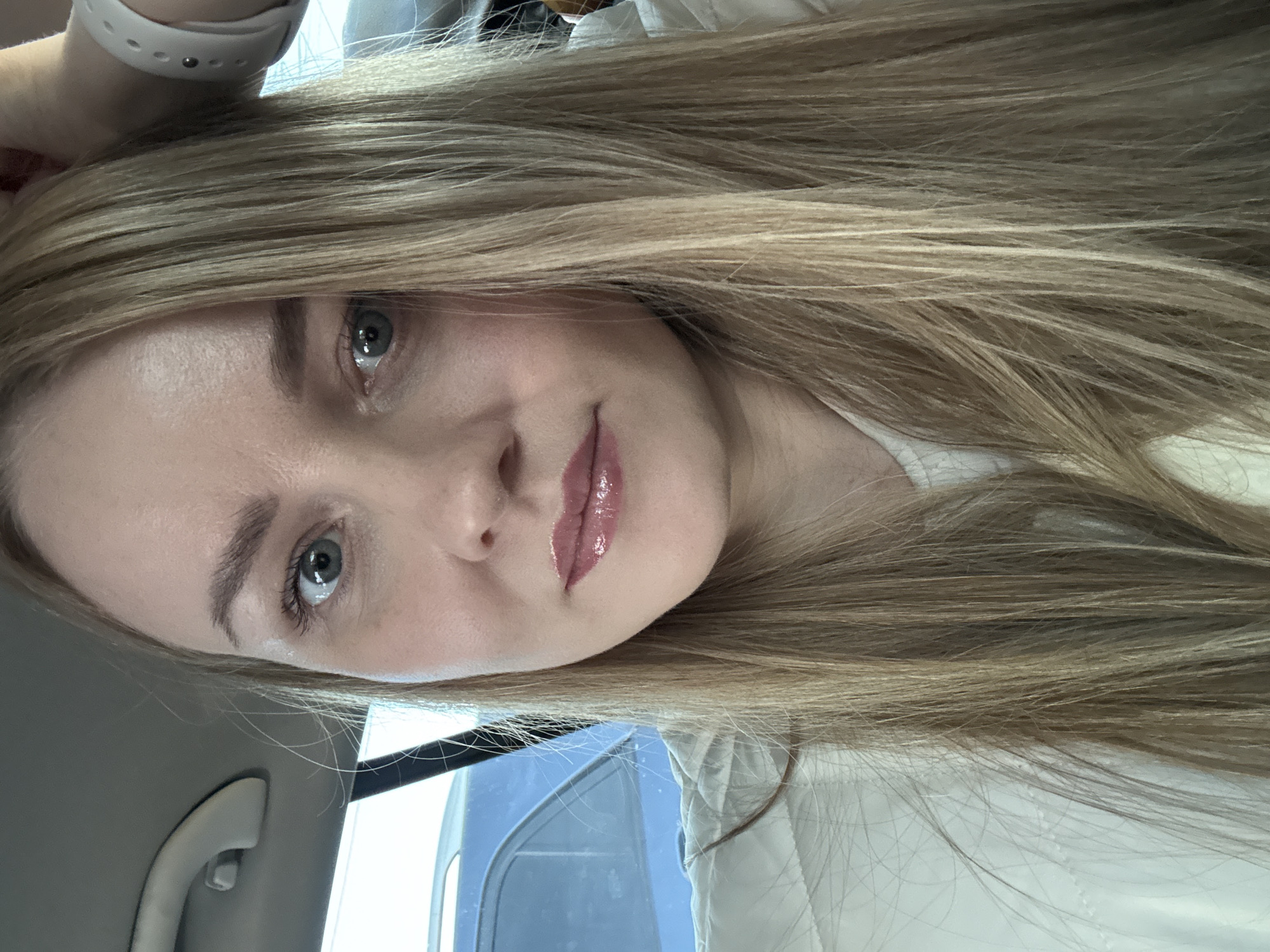 A close-up selfie of a young woman with long, blonde hair and light eyes. She is wearing a white top and what appears to be a smartwatch on her left wrist. Her makeup includes glossy lipstick. The photo appears to have been taken inside a vehicle, based on the window and interior trim visible in the background. The location is unknown and cannot be determined.