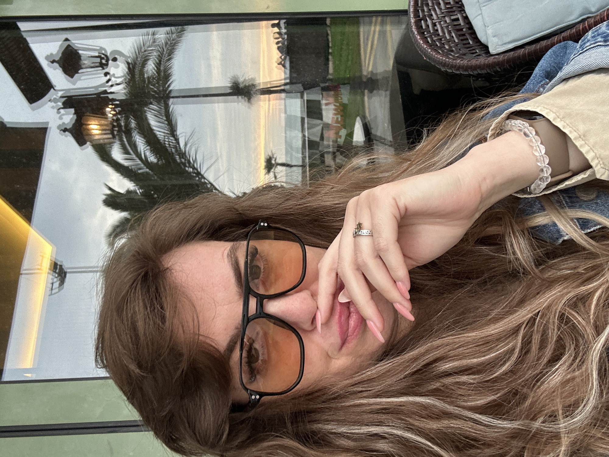 A woman with long, wavy blonde hair and orange-tinted glasses is seen close-up. Her fingers, adorned with long pink nails, rest near her face. She wears a ring and a beaded bracelet. A reflection in the window behind her shows palm trees, streetlights, and what appears to be part of the Batumi cityscape at sunset, including a glimpse of a road and surrounding greenery. A rattan chair is visible in the upper right. The setting implies an indoor location, possibly a restaurant or cafe, in Batumi, Georgia.