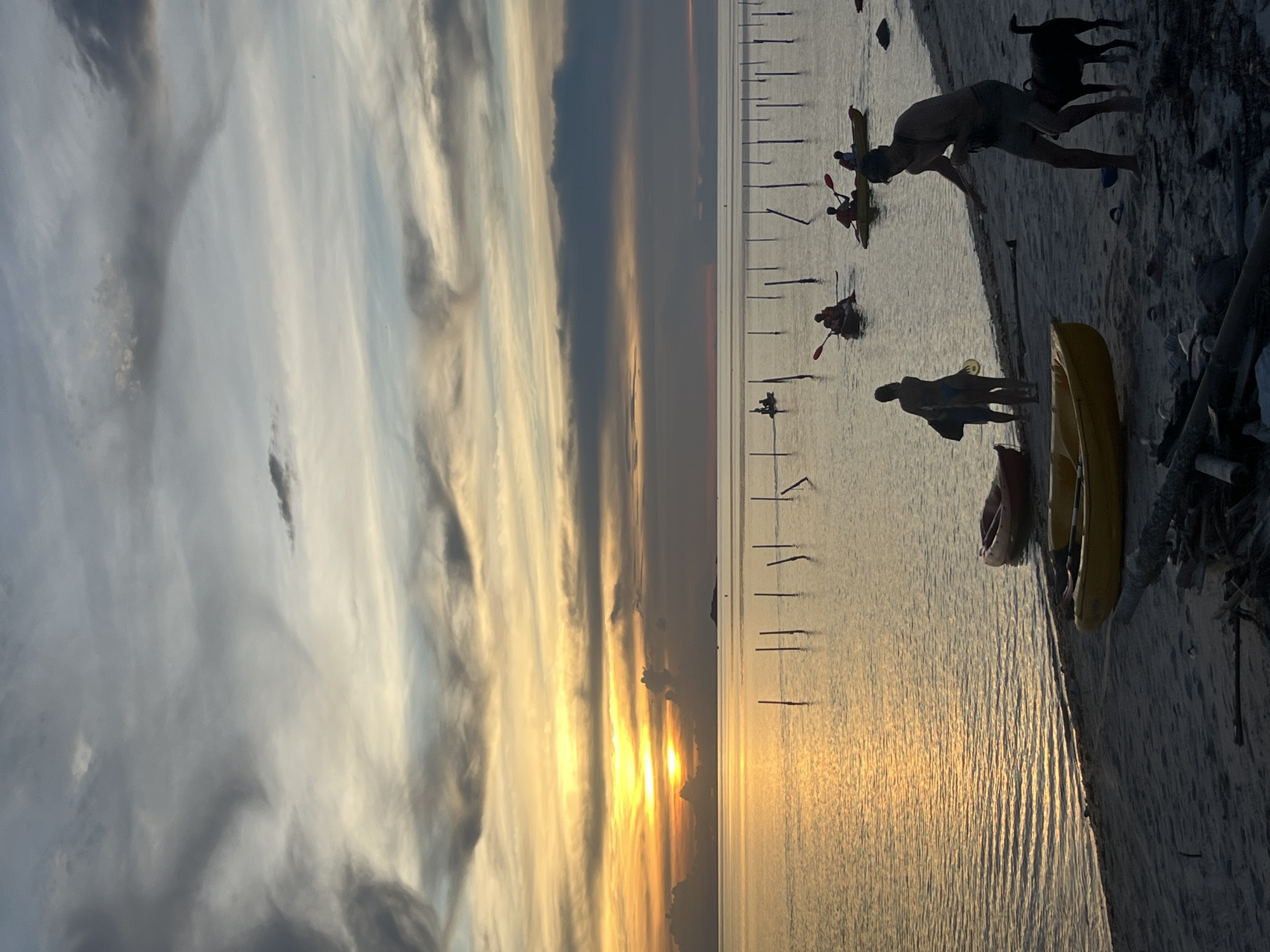 The image depicts a serene beach scene at sunset in an unspecified location. The sky is filled with dramatic, swirling clouds in shades of gray and orange, with the sun casting a golden glow on the calm water. The ocean reflects the warm hues of the sunset, creating a shimmering effect. In the foreground, a sandy beach is littered with debris, including driftwood and scattered items. Two kayaks, one yellow and one red, are beached near the water's edge.
Several people are present in the scene. On the beach, a man in shorts and a tank top is standing with a dog. He appears to be interacting with the dog, which is looking up at him. Beside him, two figures, possibly a couple, are silhouetted against the water. Another figure stands further down the beach, carrying a bag.
In the water, several individuals are kayaking. Three kayaks are visible, each carrying one or two people. They are paddling in the calm sea, enjoying the sunset. In the distance, a row of wooden poles stands in the water, possibly marking a boundary or part of a submerged structure. The horizon is visible, with a faint outline of land in the far distance. The overall atmosphere is peaceful and tranquil, capturing a moment of leisure and natural beauty.