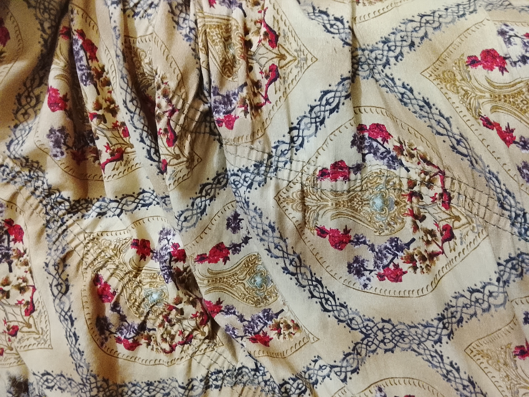 The image shows a close-up of patterned fabric. The fabric is a light beige color with a repeating pattern of square shapes. Within each square, there is a floral design featuring red and purple flowers and a decorative gold embellishment. The squares are bordered by a blue, braided-like design. The fabric is wrinkled and folded, creating shadows and highlights across the surface. The location in the city of Multan, Pakistan is not evident from the image.