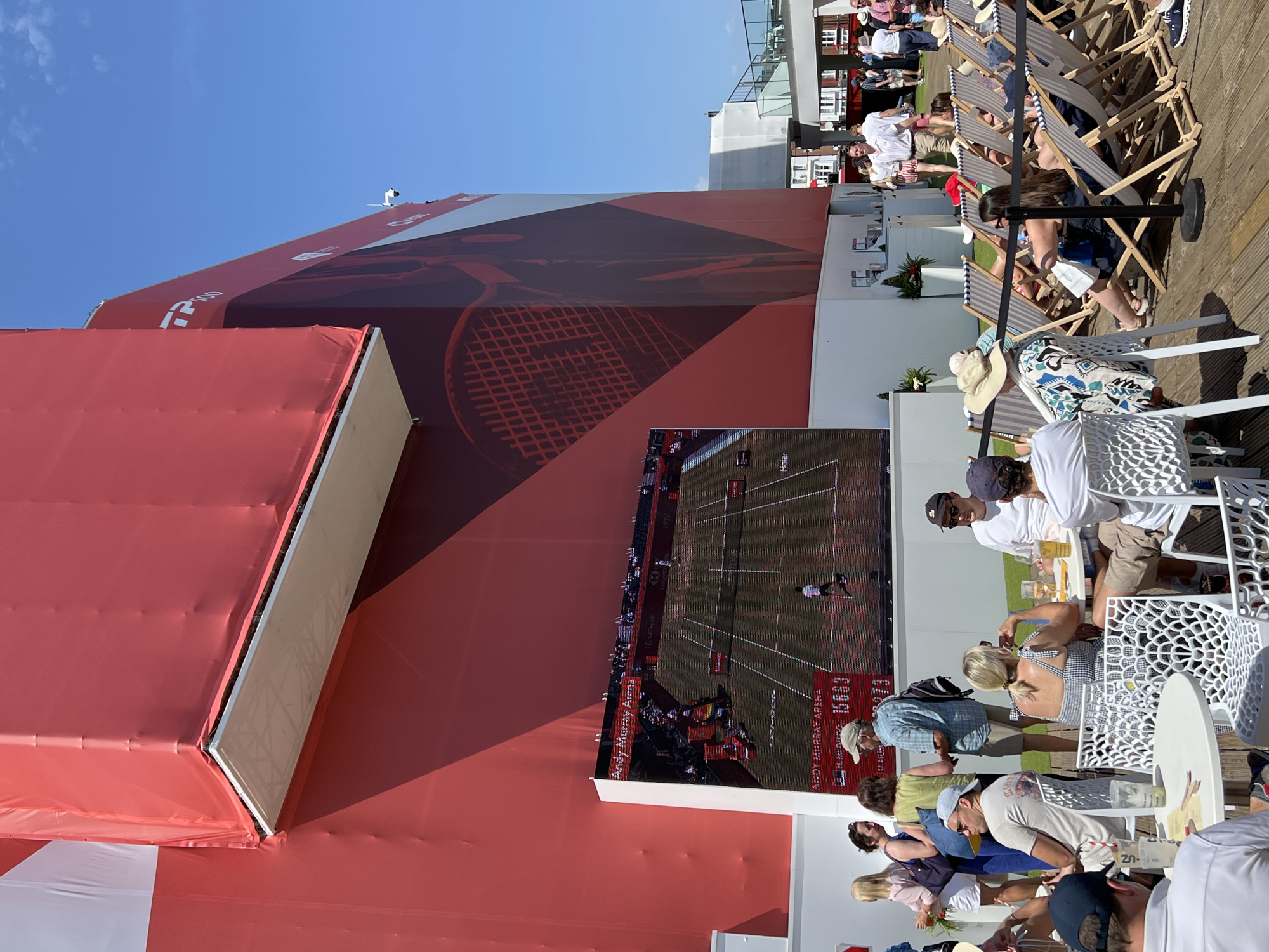 In an outdoor urban setting, a large LED screen displays a tennis match at the "Andy Murray Arena," visible to a crowd of people seated at tables and on lounge chairs. The screen shows two players on a clay court, with scores and sponsor logos. The arena is covered in a large red banner with geometric designs and a graphic of a tennis racket. In the foreground, several groups of people are casually dressed, some holding drinks and engaging in conversation, while others watch the match. The environment appears to be a sunny day with a clear blue sky. The overall scene suggests a lively event, possibly a sporting tournament, with spectators enjoying the game in a relaxed atmosphere.