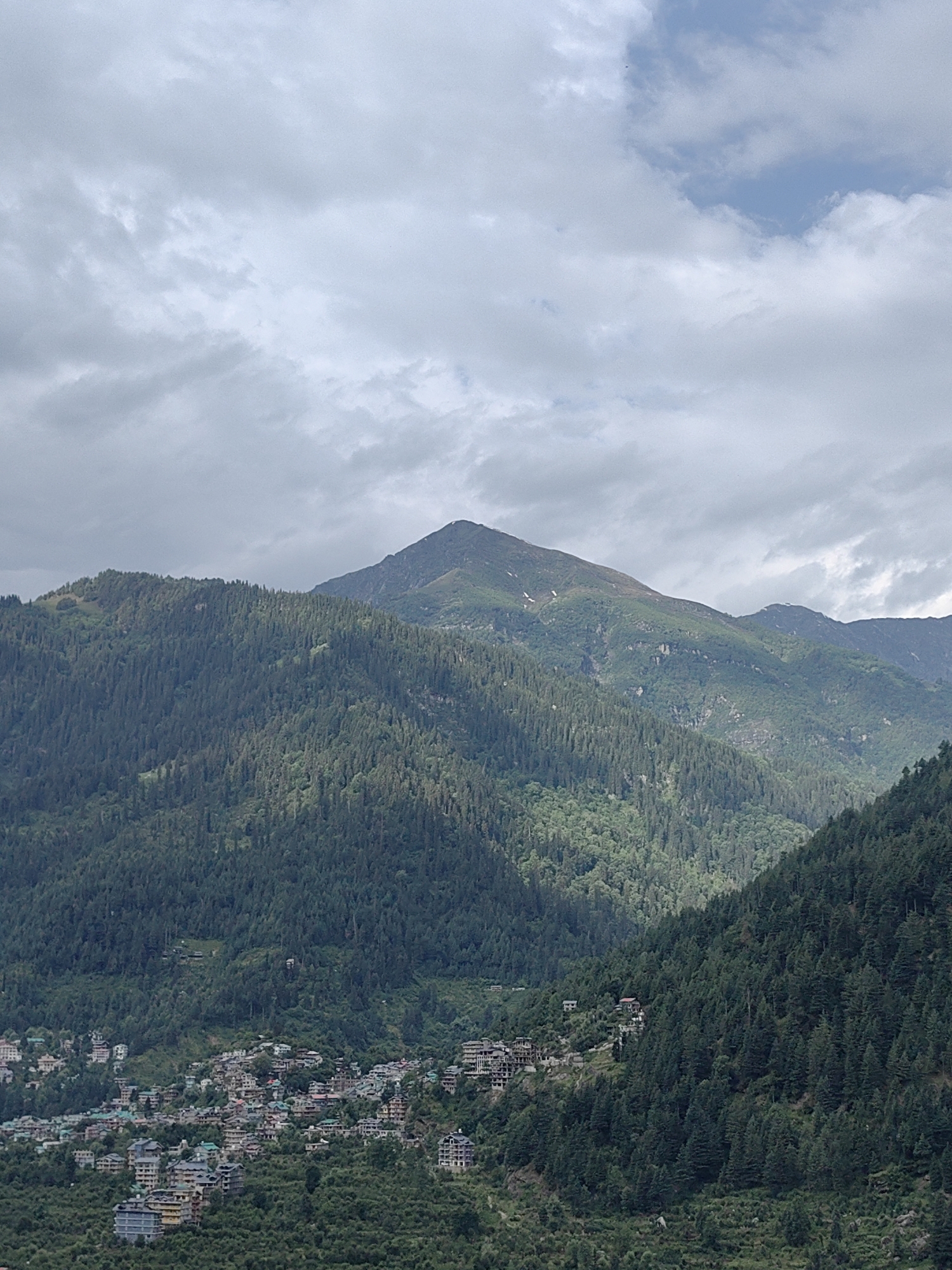 This image captures a sweeping vista of the mountainous region surrounding Vashisht, India. The foreground is dominated by densely forested mountainsides, a tapestry of dark green coniferous trees stretching across the slopes. Nestled into the lower slopes of the mountains is the town of Vashisht, a collection of buildings with varying architectural styles, many featuring pitched roofs. The town appears to be built on a hillside, with structures cascading down the terrain.
In the mid-ground, the mountains continue their ascent, with patches of lighter green vegetation and rocky outcrops visible amidst the dense forestation. The peak of a prominent mountain stands tall in the background, its summit partially obscured by clouds. The sky above is a mix of gray and white clouds, with glimpses of blue peeking through, suggesting a partly cloudy day. The overall impression is one of a serene, natural landscape punctuated by human habitation. There are no people or animals clearly visible in the image.