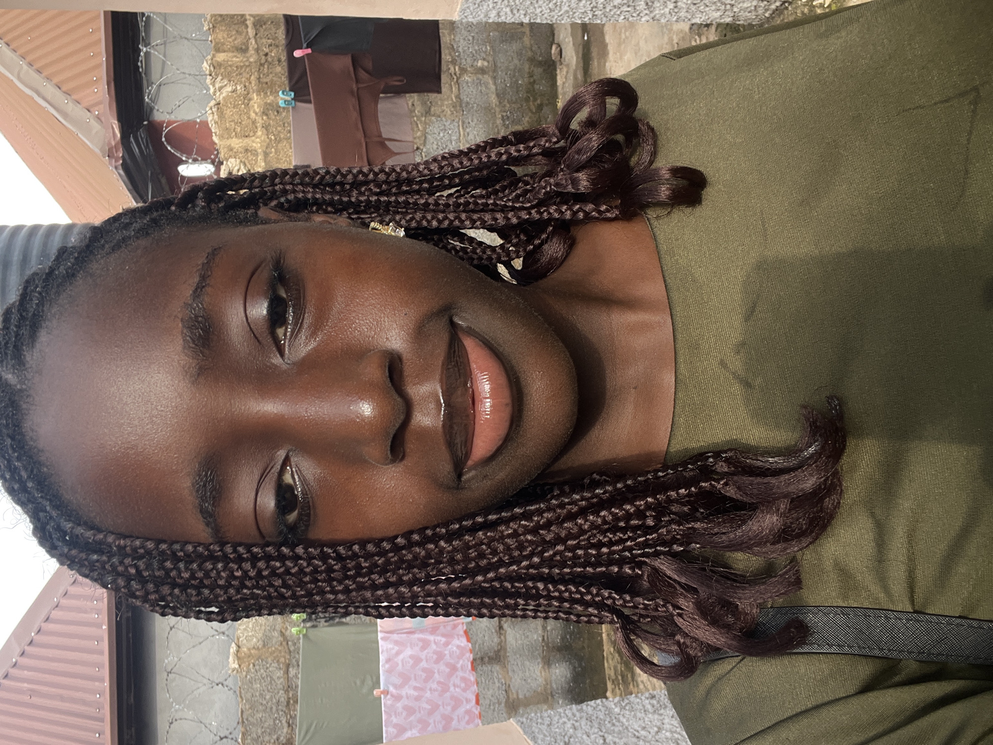 A portrait-style photograph, taken in Abuja, Nigeria, captures a young Black woman with an olive green top and dark, neatly braided hair. She is positioned slightly to the right of the frame, looking directly towards the viewer with a gentle, closed-mouth smile. Her lips appear glossy, and her skin has a luminous quality. Her hair is styled in numerous small braids (box braids), which cascade over her shoulders and chest, with the ends forming soft curls.
The setting appears to be outdoors during daytime, indicated by the bright, natural lighting. The background reveals elements suggestive of a residential or semi-urban environment. To her left, a brown corrugated metal roof is visible. Behind her, a rustic stone or concrete block wall can be seen, partially obscured by a section of barbed wire fencing. Further in the background, some white and pink fabric, possibly laundry, is hanging. A black strap, likely from a bag, is visible over her right shoulder. There is no visible text in the image.
