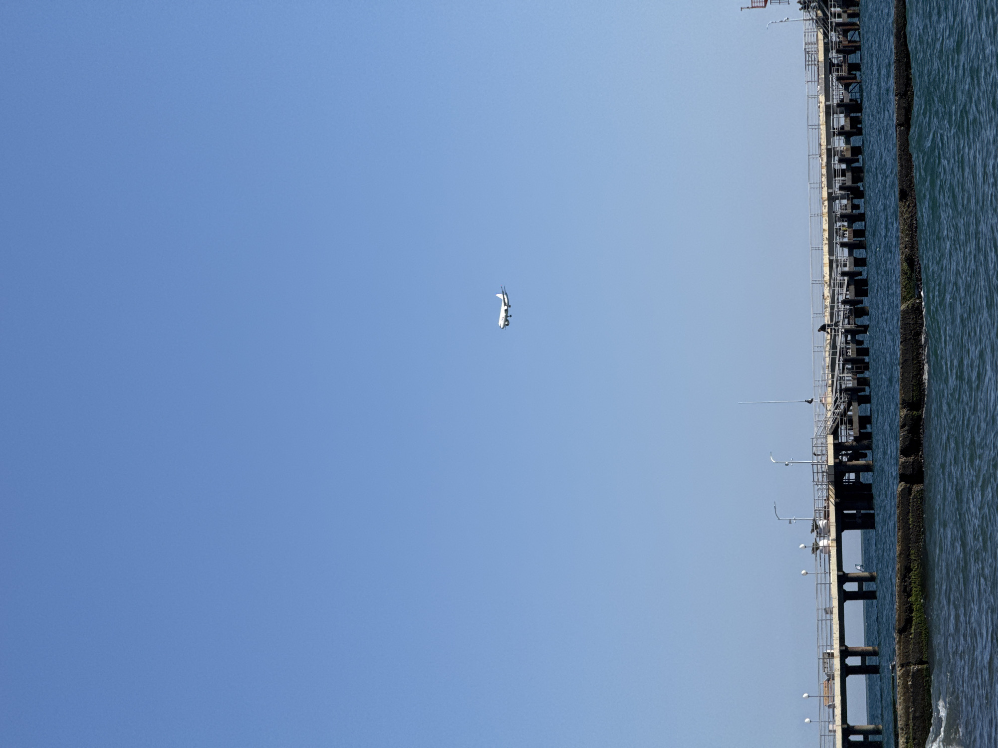 The image captures a clear, bright day over a coastal area in Sochi, Russia. The majority of the frame is dominated by a vast, cloudless blue sky, indicating excellent weather. High in the upper-left section of the sky, a white airplane, likely a commercial jet, is visible in flight.
On the right side of the image, a long pier or jetty extends into the water. This substantial structure is built on numerous dark pillars, supporting a walkway equipped with metal railings. Several white, spherical lampposts or light fixtures are spaced along the pier. Closer inspection reveals a few small, indistinct shapes on the pier, possibly birds such as seagulls, perched on the railings or structure.
Adjacent to the pier, the sea exhibits noticeable waves and ripples, suggesting movement in the dark blue-green water. The sunlight illuminates the scene, highlighting the contrast between the clear sky, the structured pier, and the textured surface of the water. The overall impression is one of a peaceful, sunny coastal environment, with an airplane traversing the clear skies above. No visible text is present in the image.
