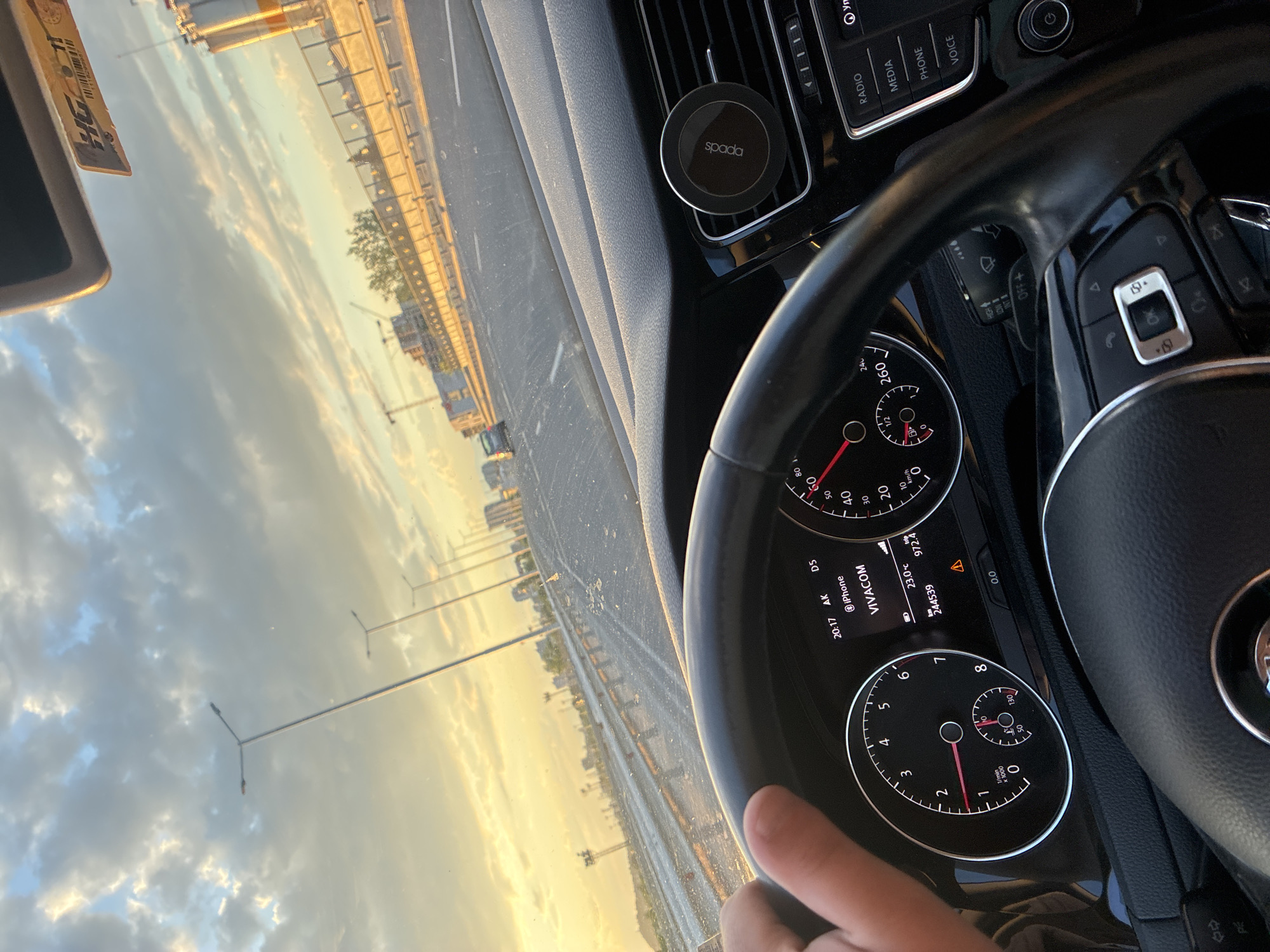 The photograph was taken from inside a vehicle, focusing on the dashboard, steering wheel, and the road ahead. The vehicle's speedometer shows a speed of 0 km/h. The dashboard display indicates the time as 20:17, a temperature of 23.0°C, and a total mileage of 244,539. The Spada car phone holder is attached to the vents on the dashboard. Through the windshield, one can see a highway, overhead electrical lines, buildings, and a cloudy sky.
