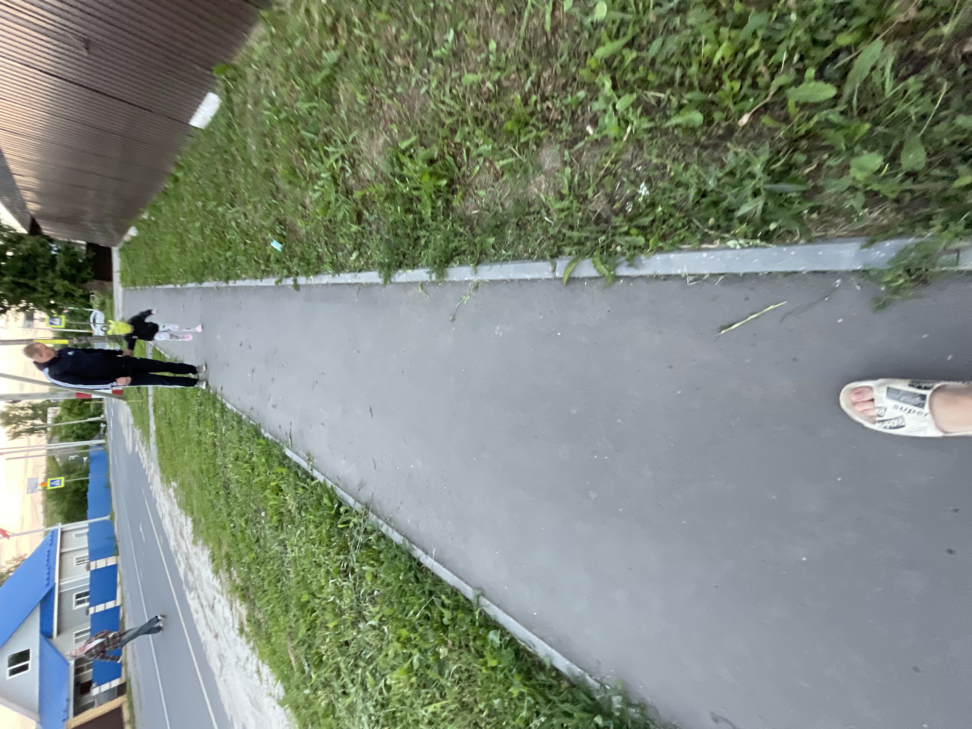 A young child wearing a black jacket, green hat, and patterned pants runs on a sidewalk next to a man in a black tracksuit. The man is holding the child's hand. The sidewalk is made of grey asphalt and bordered by grass. In the background, there is a brown metal fence, trees, and buildings, including a house with a blue roof. A road is visible to the left, with a person on a skateboard in the distance. The scene appears to be in an outdoor, urban setting in Красные Баки, Russia.