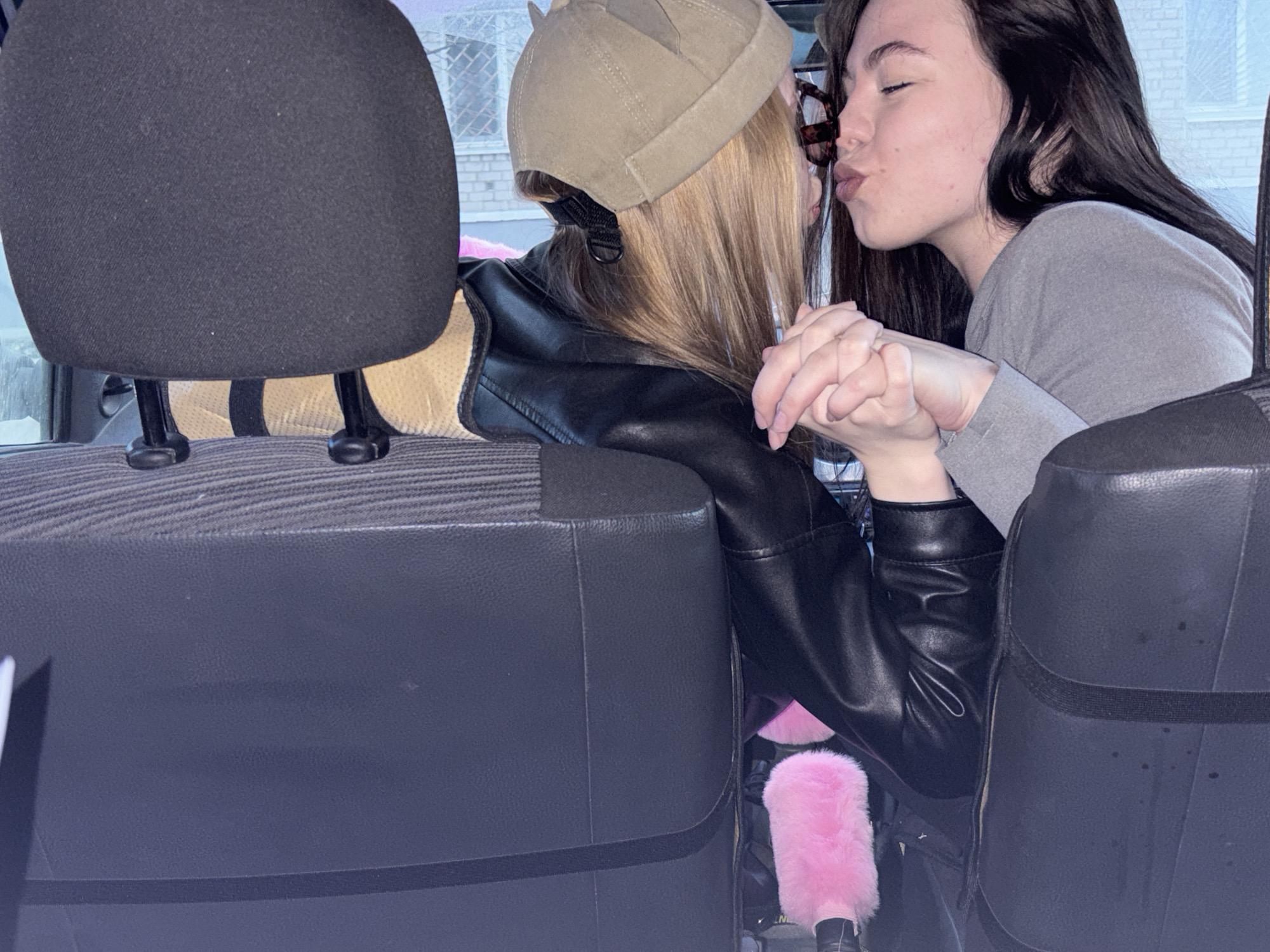 Two women are seated inside a car in what appears to be Perm, Russia, as indicated by the view through the car window showing a brick building, possibly a residential or commercial structure. The woman on the left wears a tan cap and a black leather jacket. The woman on the right has dark hair and wears a gray long-sleeved shirt. Both women are positioned so that their faces are near one another, with their lips close, possibly kissing. Their hands are clasped together. Pink furry objects are attached to the car's interior near the women. The interior of the car is visible, including the headrests and the seat upholstery.