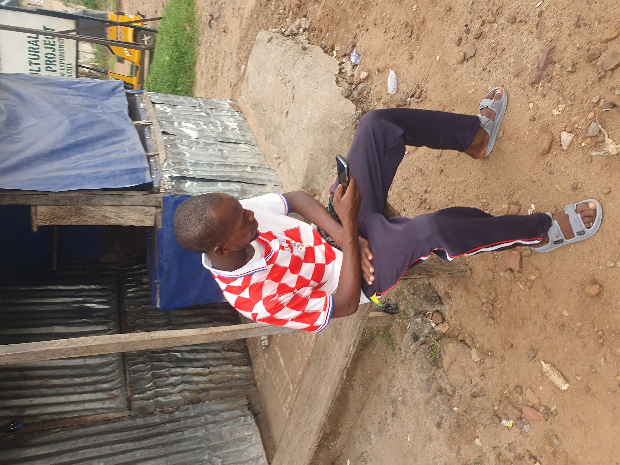A young Black man is sitting on a wooden plank bench outside a structure made of corrugated metal sheets. He is wearing a red and white checkered shirt and dark pants with white and red stripes down the sides. He is holding a black smartphone and looking down at it. His feet are bare except for grey sandals. The ground is dirt and scattered with small rocks and debris, including a discarded plastic bottle. In the background, a blue tarp is draped over part of the structure, and a sign is visible with the words "CULTURAL PROJECT" and "EXPRESSWAY STATE". A yellow tricycle is parked behind the sign. This scene is likely located in Lokoja, Nigeria, given the context of the sign mentioning "EXPRESSWAY STATE," which is a common road designation in Nigeria.