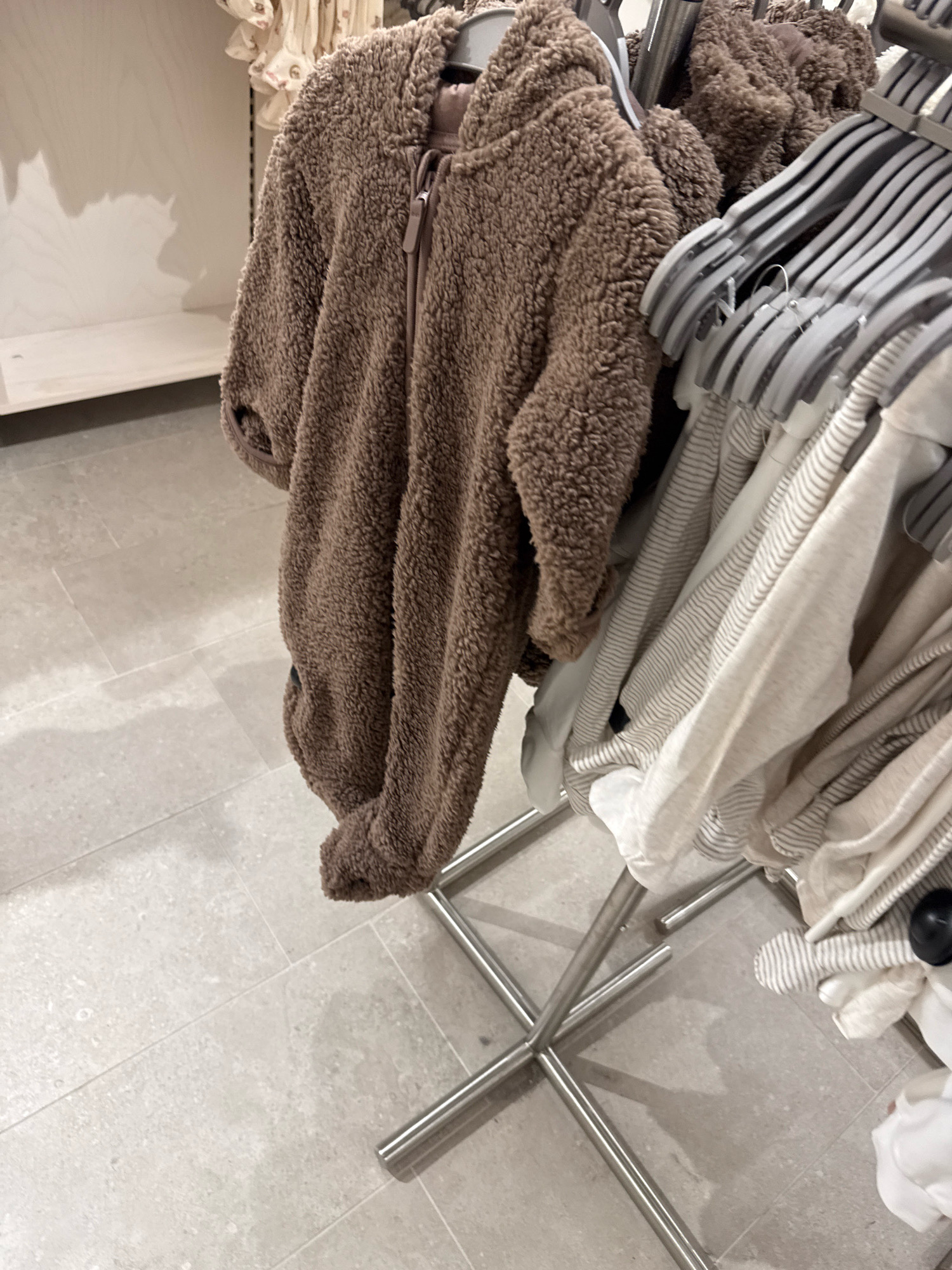 A brown, fuzzy onesie is hanging on a clothing rack in what appears to be a retail store. The onesie is a one-piece garment with a zipper closure and a hood. It is made of a soft, plush material. Other clothing items, including striped shirts and lighter colored garments, are also visible on the rack. The floor is tiled with light gray tiles. A shelf is visible in the background. The scene is well-lit, likely from overhead store lighting. There are no people or animals present in the image. The location is a clothing store in Tallinn, Estonia.