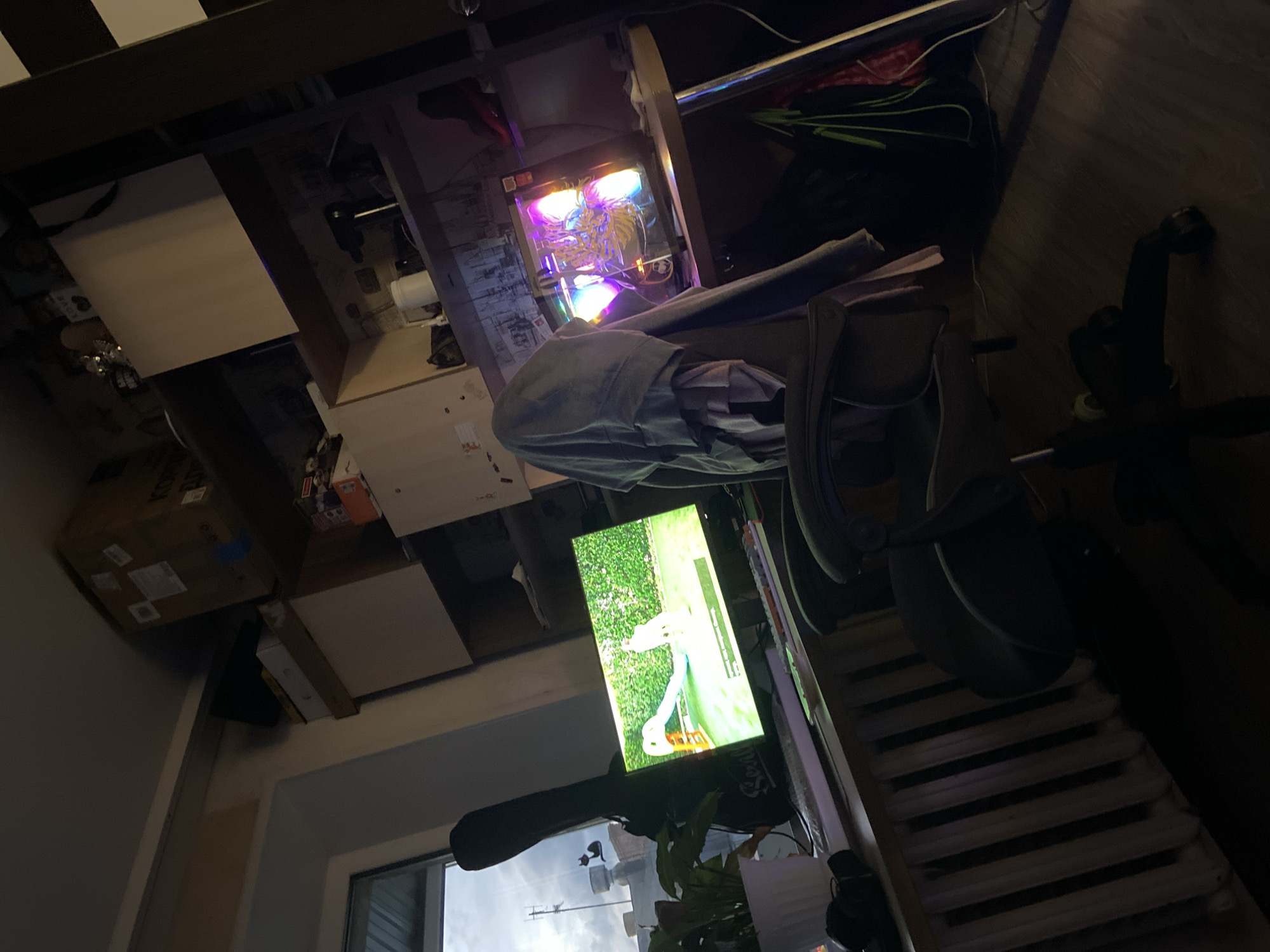 The image depicts a cluttered room, likely a home office or gaming setup, with a desk area prominently featuring a computer and a monitor. The computer tower is illuminated with colorful RGB lighting, suggesting it is powered on and possibly in use. On the desk, a monitor displays a video of a goose on a golf course. Various objects are scattered around, including boxes, what appears to be a gym bike, and some clothing or fabric draped over a chair. The lighting in the room is dim, with the computer's lights and the monitor being the main sources of illumination, indicating it is likely evening or night. The overall impression is one of a lived-in space filled with electronics and personal belongings. There is no visible text in the image.