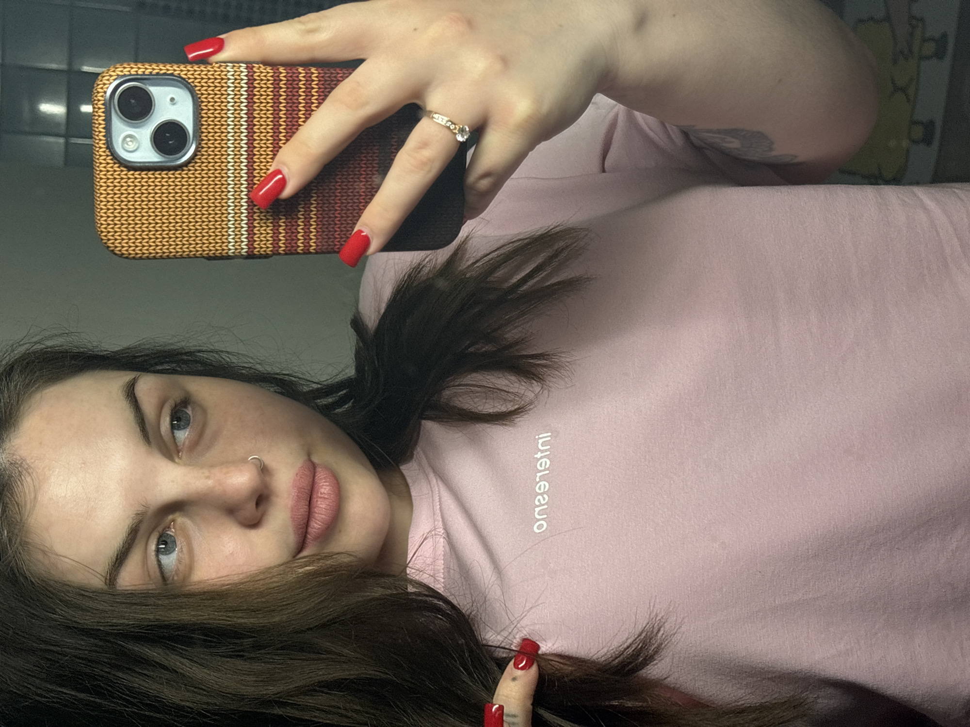 A woman is taking a selfie in what appears to be a bathroom. She is holding a smartphone with a knitted-patterned case in her right hand, showing red nail polish on her fingernails and a ring on her ring finger. She is wearing a light pink t-shirt, with the word "interismo" printed in reverse on the front. Her hair is dark brown, and she has a nose piercing. The background appears to be a bathroom with dark-colored tiles visible. The location in the city of Chornomorskoe, Ukraine, cannot be determined from the image.