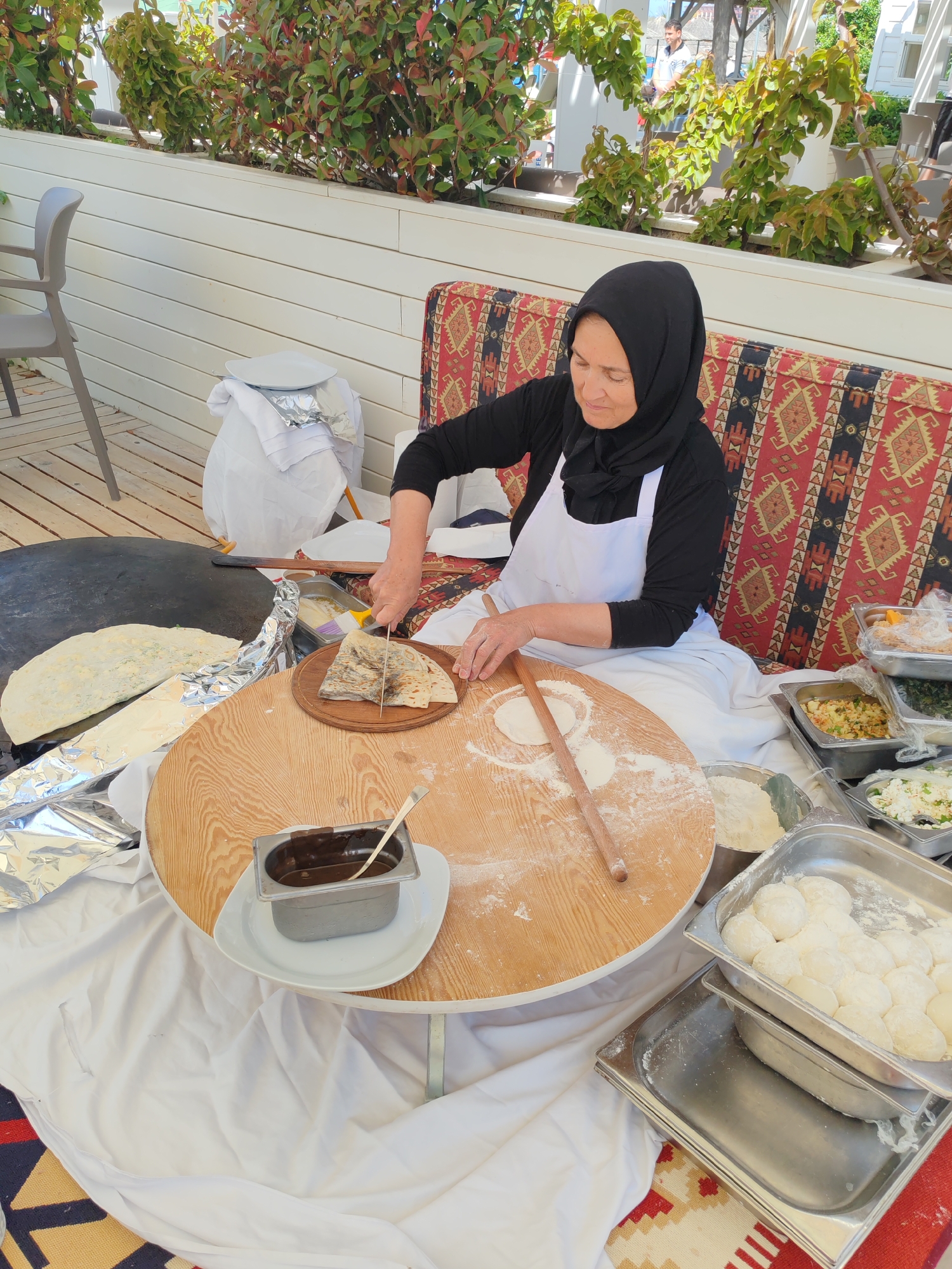 An outdoor scene in Alanya, Turkey, shows a woman preparing food at a makeshift cooking station. The woman, wearing a black hijab and a white apron over a black long-sleeved shirt, sits on a cushion with a patterned red and black design. She is cutting a flatbread on a small wooden board with a knife. A round, wooden table in front of her is dusted with flour, and a rolling pin lies on the surface.
To the left of the table is a large, flat griddle with cooked flatbreads, some covered in foil. A small metal container filled with a dark liquid sits on a white plate near the table's edge. To the right, stainless steel trays hold ingredients and dough balls.
The cooking station is set against a white, slatted wall adorned with green plants. A gray chair is visible in the upper-left corner. People can be seen in the distant background. A white cloth drapes over the table and the floor.