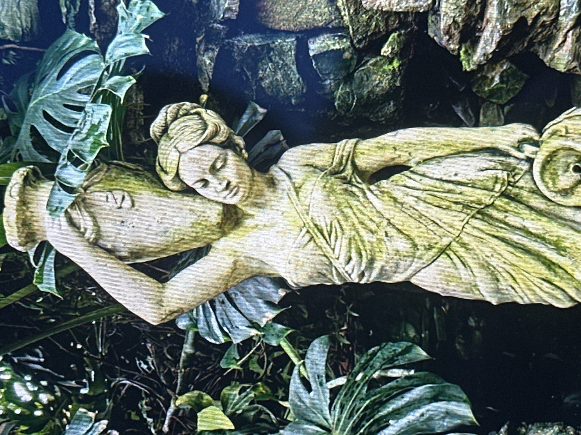 A statue of a woman, weathered to a pale green and yellow, stands in a garden. She is depicted in a classical style, wearing a draped garment and has her hair pulled up in a bun. Her right arm is raised, supporting a large amphora or vase on her shoulder, while her left hand holds a small bowl or censer at her side. Lush green foliage, including large-leafed plants and vines, surrounds the statue, with rough, moss-covered rocks in the background. The lighting suggests an outdoor setting, possibly during the day. The statue appears to be the focal point of a garden or decorative display.