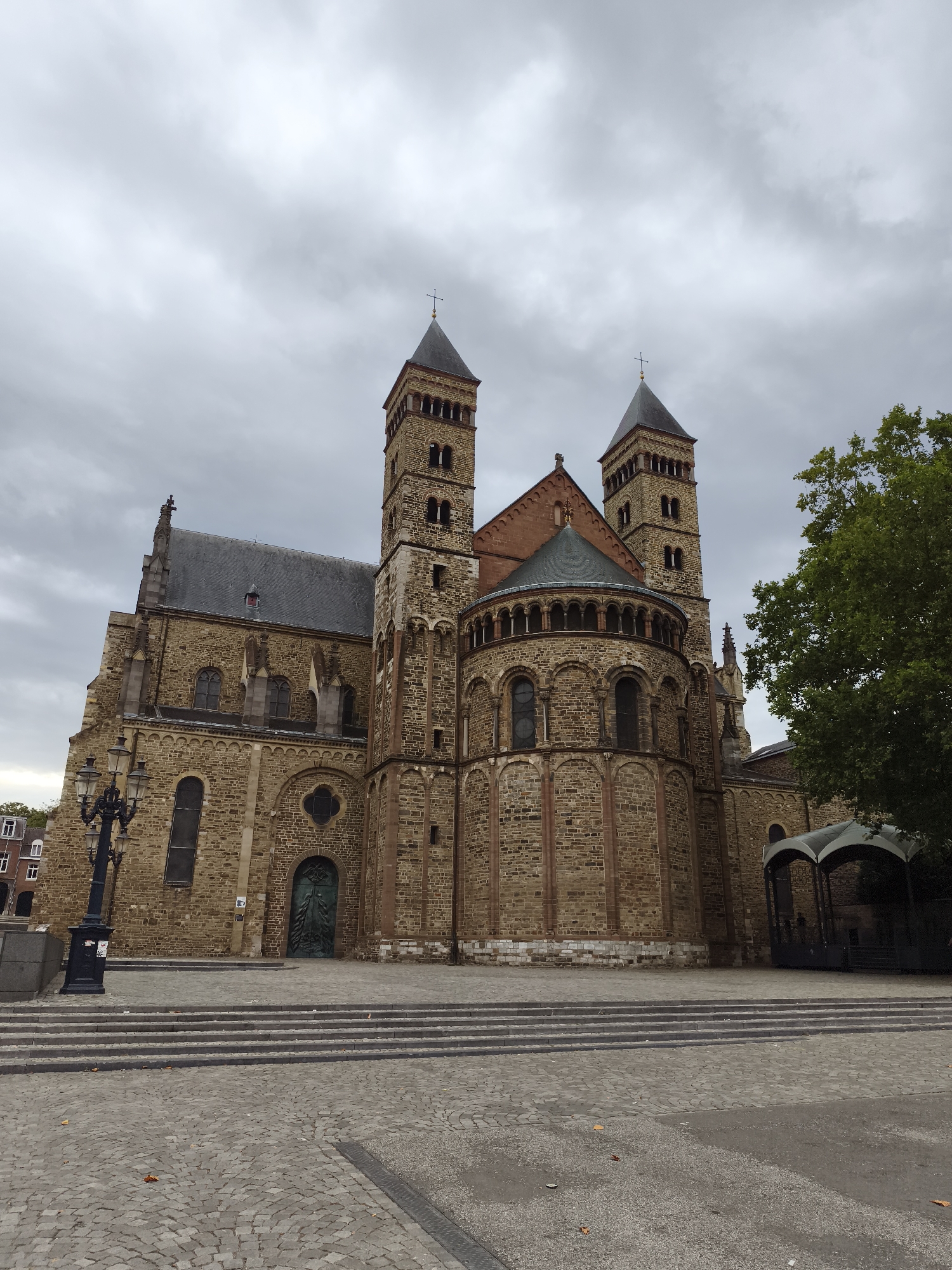 This is a full-frame, eye-level, outdoor, medium shot of the Basilica of Saint Servatius in Maastricht, Netherlands. The sky is overcast and grey. The foreground is a cobblestone plaza with several wide steps leading up to the basilica. To the left of the steps is a black street lamp with multiple lights. The basilica is constructed of rough, brown stone with red accents. It features two prominent square towers with pointed roofs, and a large, rounded apse at the rear. The main entrance door is a large, dark green, arched door with ornate detailing. A portion of a mature green tree is visible on the right side of the frame, extending from the top right corner down to the middle right. The overall scene is solemn and architectural.