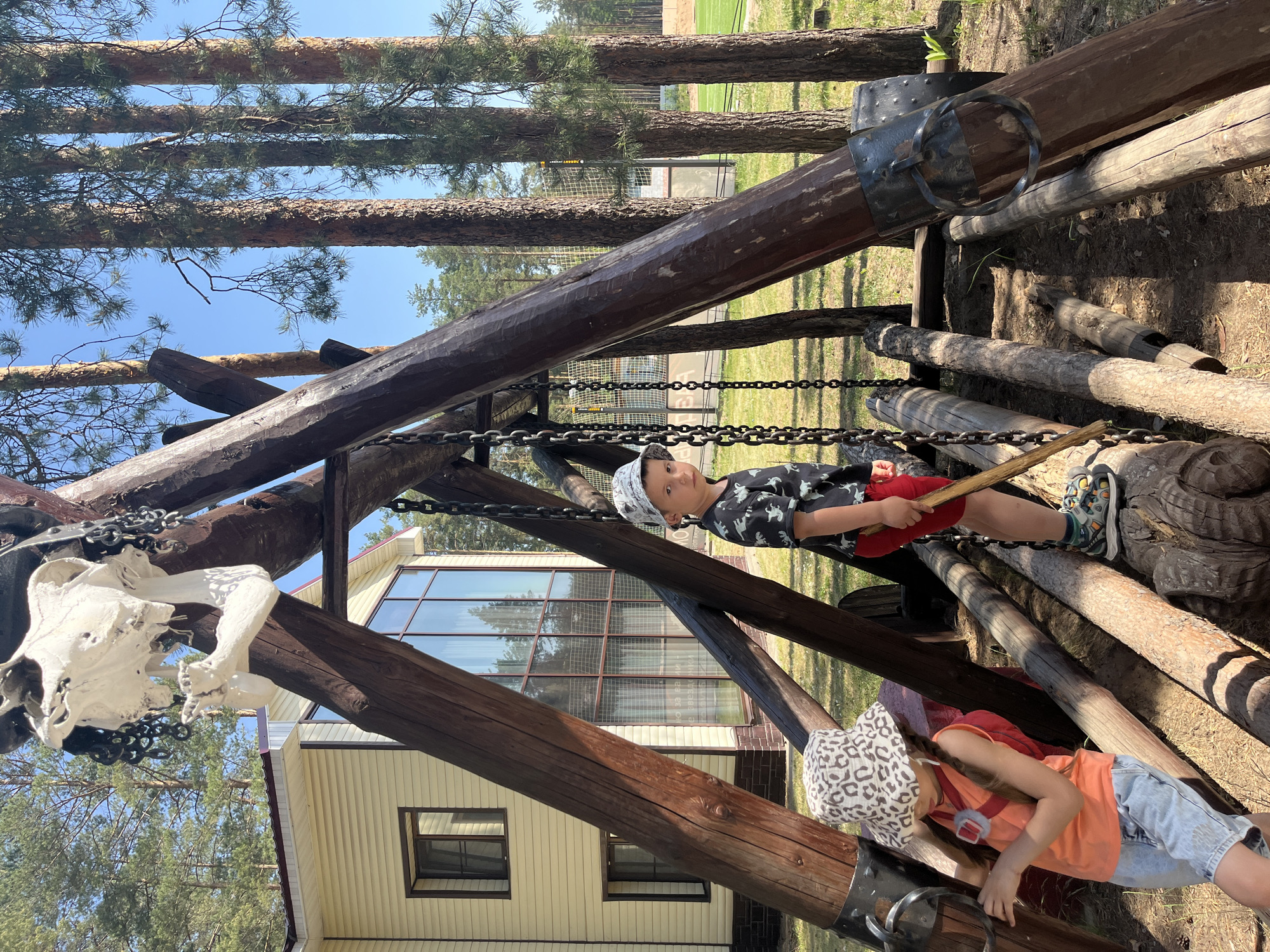 Two children play on a wooden playground structure. The boy is in the center holding a brown stick, while the girl is at the bottom right. The structure has logs, chains, and a skull hanging from it. A two-story building is in the background with windows and light yellow siding. Trees are in the background and blue sky is above. The location is Chaykovsky, Russia.