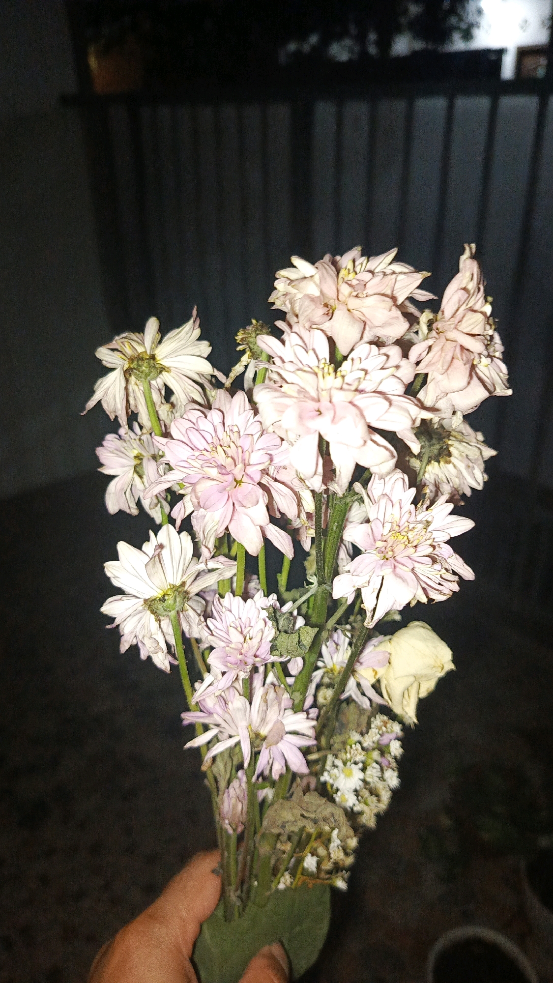 A hand holds a bouquet of light pink and white flowers, some of which appear wilted or dry. The background is dark and features a blurred vertical pattern, possibly a fence or blinds. The scene is dimly lit. The image was taken in Babadan, Indonesia.