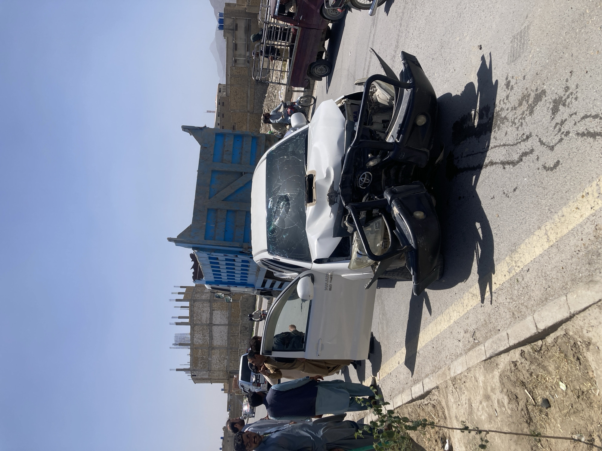 The image shows a wrecked white Toyota Hilux pickup truck on a paved road. The front of the truck is heavily damaged, with a smashed windshield. The driver-side door is open, revealing a portion of the interior. Several people are standing near the truck, some appearing to be inspecting the damage. In the background, there is a blue and white truck, a maroon truck, and some motorcycles. The location appears to be an urban area with buildings and construction in the distance. Based on the urban setting and context clues, the location is likely Quetta City Tehsil, Pakistan.