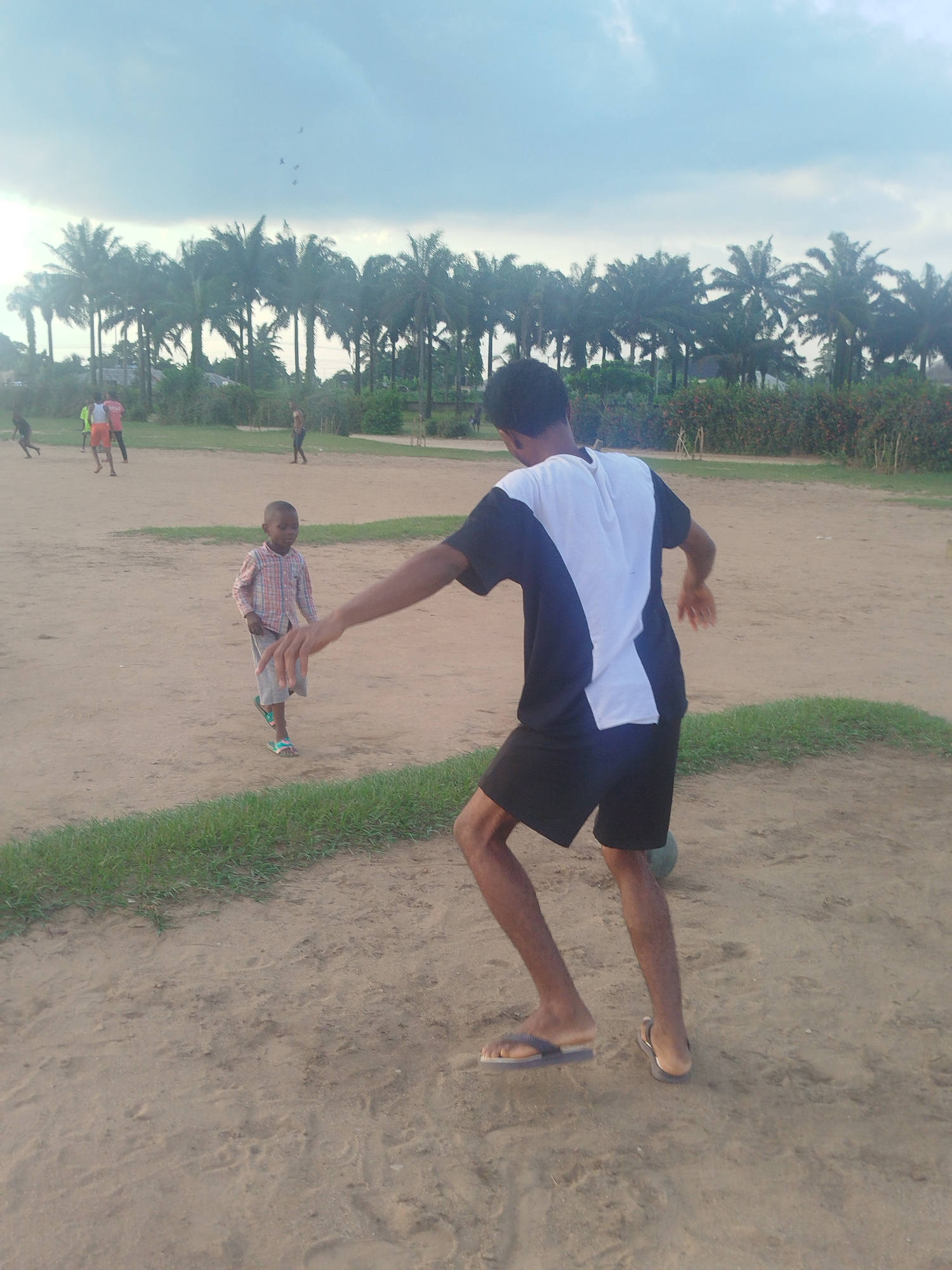 A medium shot shows a sandy, dirt-covered field in Ikot Mbonde, Nigeria. In the foreground, a young man with dark hair and skin is wearing black shorts and a two-tone black and white shirt, playing with a soccer ball at his feet. He's wearing flip-flops and has one arm extended forward. A young boy in a checkered shirt and shorts stands in front of him, appearing to watch. In the background, several other figures are engaged in a soccer game. Beyond the soccer field, there is a row of tall palm trees. The sky is overcast with a hint of dusk.