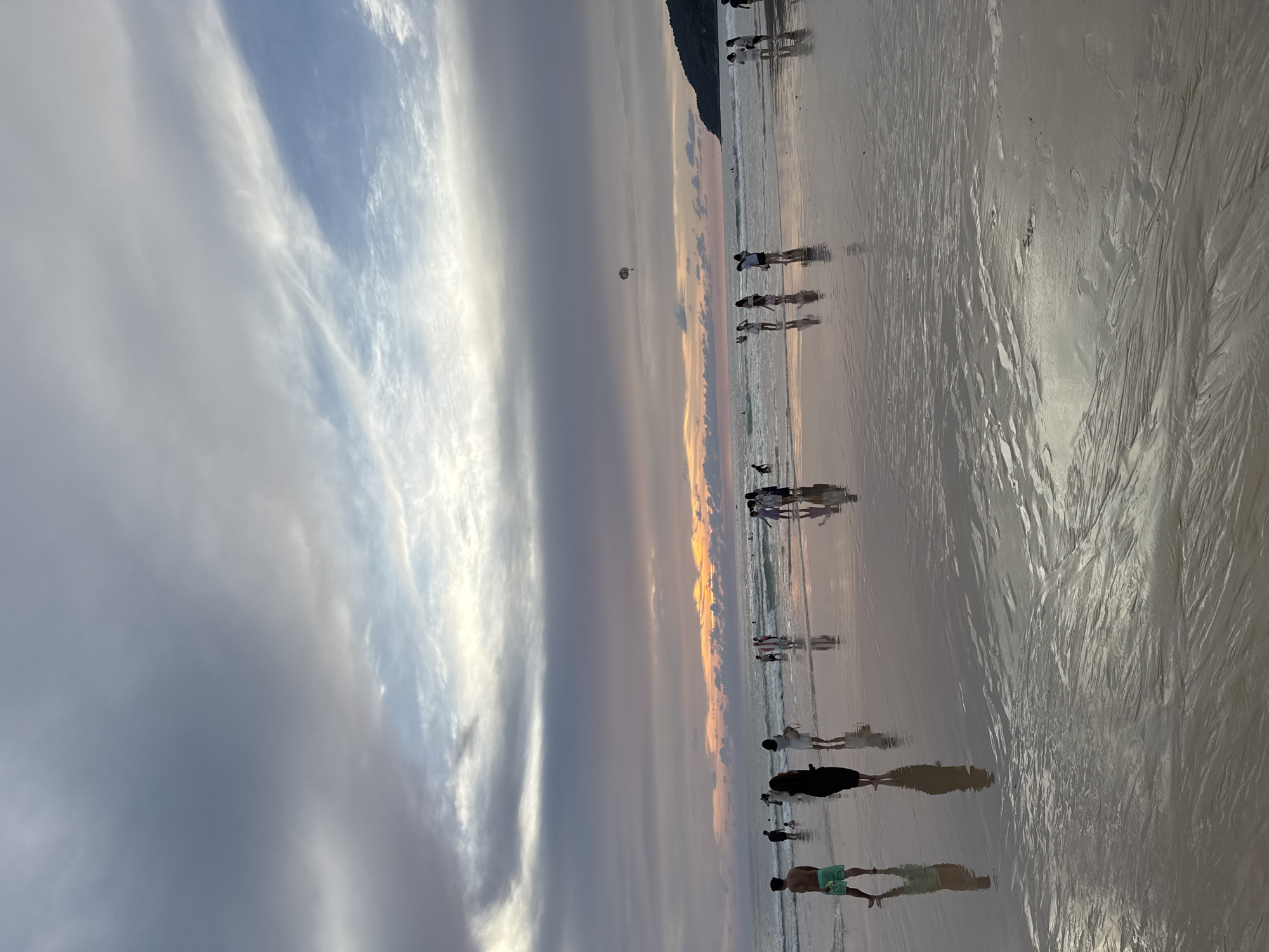 The image depicts a beach scene at sunset in Karon, Thailand. The sky is filled with a dramatic display of clouds, transitioning from dark grey on the left to lighter blues and soft pinks and oranges near the horizon, indicating the setting sun. Several people are visible on the wet sand and in the shallow water.
In the foreground, a man wearing green swim trunks stands facing away from the camera, his reflection visible on the wet sand. To his left, a woman in a black dress also stands, her reflection mirrored on the beach. Further back, a group of individuals are walking along the water's edge and wading in the sea. Some are silhouetted against the bright horizon, while others are more clearly discernible.
The ocean has moderate waves rolling onto the shore, and the wet sand reflects the sky and the figures on the beach. In the distance, a parasailer is visible in the sky, and a dark, tree-covered hill or mountain forms the far right edge of the landscape. The overall atmosphere is tranquil, characteristic of a beach at dusk.