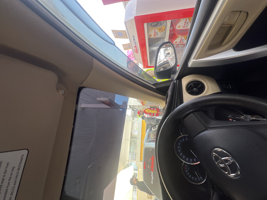 The image captures a scene from within a Toyota vehicle, identified by the steering wheel emblem, parked at a Shell fuel station in Abbottabad, Pakistan. The perspective is from the driver's seat, looking forward through the windshield and towards the passenger side during daylight hours.
Inside the vehicle, the black steering wheel, a dashboard displaying a speedometer marked to 220 km/h, and an ignition with keys are visible. A beige-colored air vent is present on the passenger side dashboard, and an overhead sun visor contains a printed warning regarding child seat safety. A dark weather deflector is affixed to the passenger side window frame.
Outside, under a Shell-branded canopy, a fuel station attendant, wearing a red uniform shirt, a yellow reflective vest with Shell logos, and a red cap, is actively dispensing fuel into a silver sedan positioned to the left of the viewer's vehicle. An additional individual, dressed in a dark cap and shirt, stands near the silver sedan. To the right, outside the passenger window, a Shell display cabinet branded "Shell Helix Ultra" showcases "Shell Rimula" oil products, with multiple QR code posters on its side. A masked person is discernable in the reflection of this display cabinet. A multi-story building with yellow trim is visible in the background beyond the immediate fuel station infrastructure.
