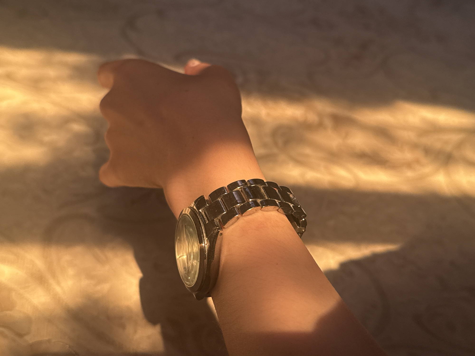 The image shows a close-up of a person's arm wearing a silver metal watch. The arm is positioned diagonally across the frame, with the watch on the wrist. The background is a textured surface, possibly a wall or fabric, illuminated by warm, golden light that casts shadows and highlights. The lighting suggests it is either sunrise or sunset. There is no discernible city or location information present in the image.