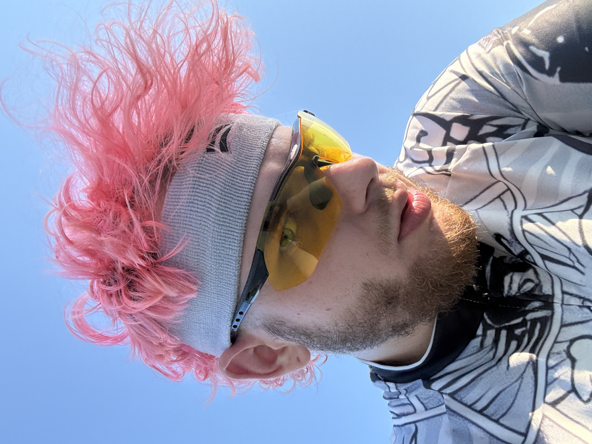A low-angle, close-up shot depicts a man with pink, frizzy hair and a light gray knitted headband. He is wearing yellow-tinted sunglasses with black frames. He has fair skin and a light brown beard. He is wearing a white shirt with a geometric black and gray pattern. The background is a clear blue sky. The location is not provided, and there is no indication the photo was taken in Đà Nẵng, Vietnam.