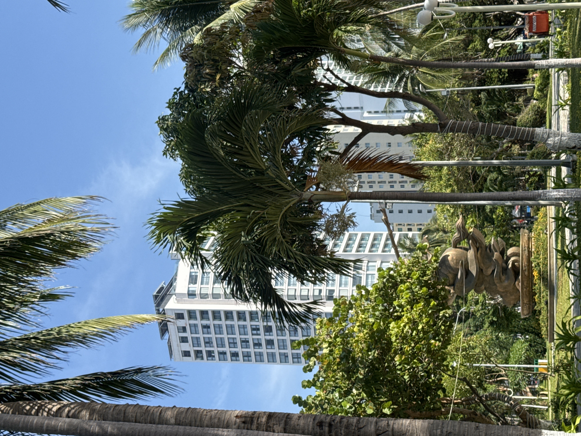 The image shows a tropical park scene in Nha Trang Ward, Vietnam. In the foreground, there is a bronze sculpture on a circular base, surrounded by lush green grass and plants. Several palm trees with rough, textured trunks are visible throughout the scene, with their fronds extending towards the bright blue sky. Behind the sculpture and trees, a modern, white high-rise building with many windows stands tall. The building appears to have a flat roof with some structures on top. In the background, more greenery and palm trees are present, along with streetlights. The overall atmosphere is bright and sunny, typical of a tropical climate.