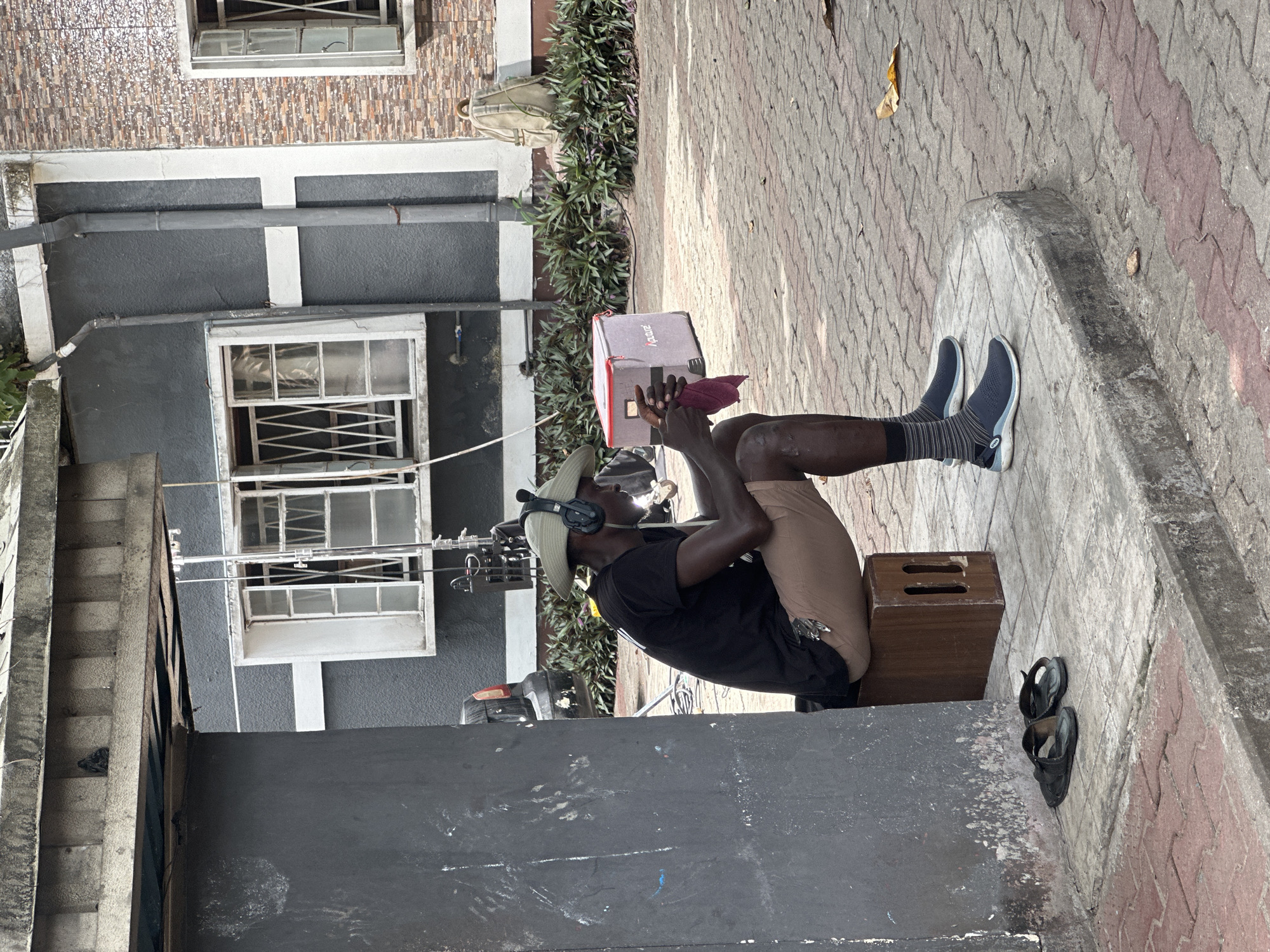 A man wearing a beige cowboy hat and headphones sits on a wooden crate on a paved surface. He is wearing a black t-shirt, khaki shorts, black socks with gray stripes, and dark blue sneakers. He holds a small red item with both hands in front of a red and gray box. Sandals lie on the ground beside the crate. In the background, a building with gray walls and white trim is visible, with several windows. The paved surface has a pattern of gray and pink bricks. The scene is located in Lagos, Nigeria.