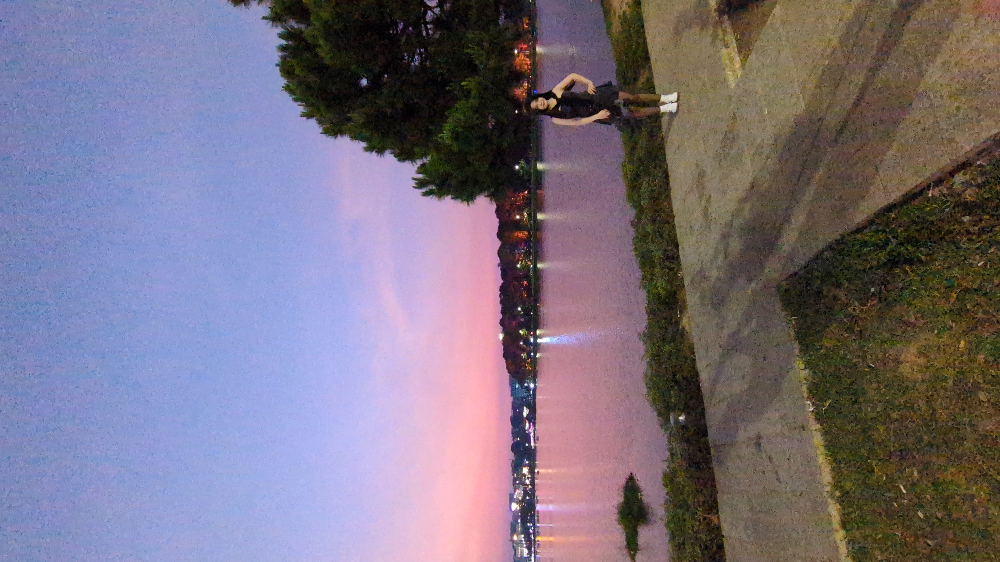 A woman in a black dress and white shoes stands on a concrete path next to a body of water reflecting lights, likely in Da Lat, Vietnam. The sky transitions from blue to pink above the water, indicating either sunrise or sunset. Trees line the water's edge, and lights from buildings or street lamps are visible in the distance. The path is bordered by grass and foliage.