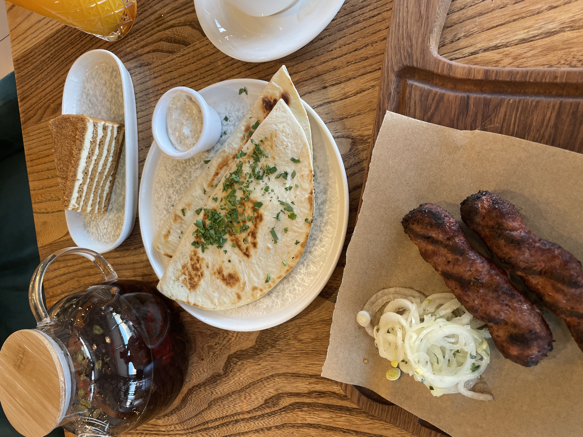 A still life composition features a meal on a wooden table. In the upper left corner, a glass teapot filled with dark liquid and herbs sits on the table. To the right of the teapot, a slice of layered honey cake is presented on a white oval platter. In the foreground, two grilled sausages rest on brown parchment paper, accompanied by a pile of thinly sliced white onions. A white dipping sauce is served in a small ramekin next to folded flatbreads, which are sprinkled with chopped green herbs. A glass of orange juice is visible on the far right. The overall scene suggests a casual dining setting.