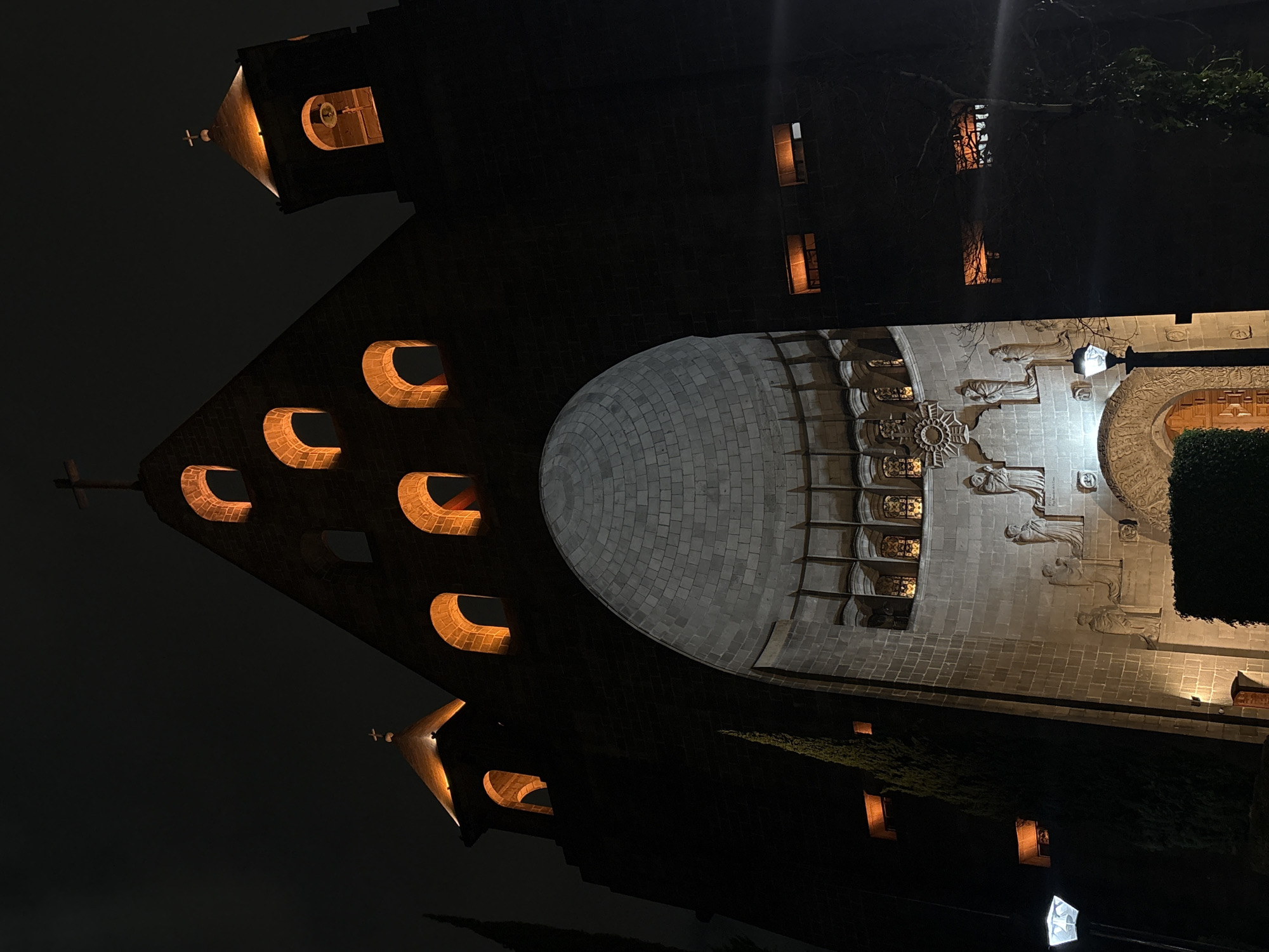The image depicts the Templo Expiatorio a Cristo Rey, located in the city of Mexico City, Mexico. The view is captured at night, with the temple illuminated by both internal and external lights, contrasting with the dark sky. The architectural details are highlighted, including the semicircular windows, the dome, and the carved statues on the exterior facade. The environment consists of the dark night sky and some greenery visible in the foreground. The image does not include people.