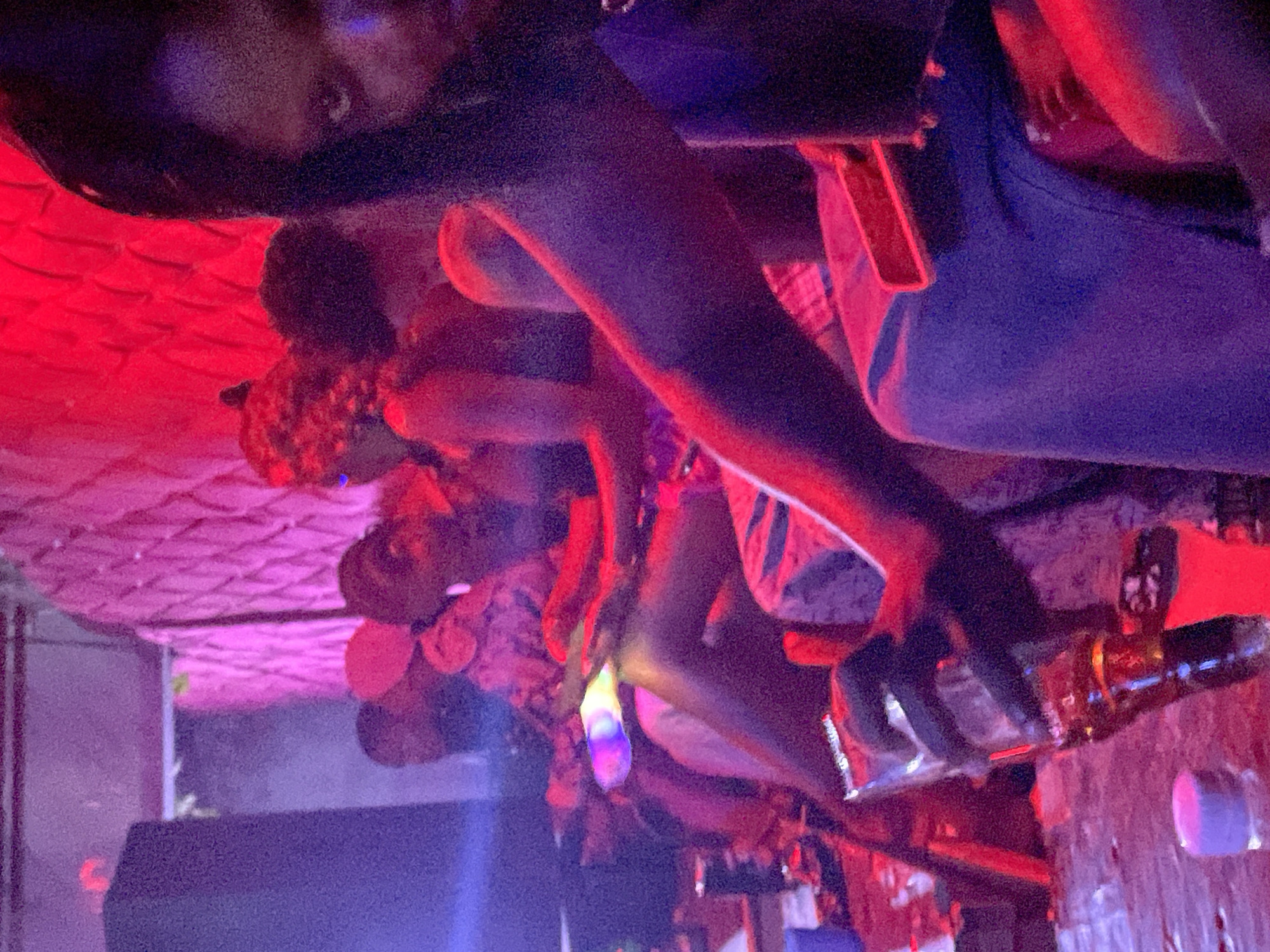 In Durumi, Nigeria, a group of people are gathered in a dimly lit area with prominent red lighting. The scene appears to be indoors, possibly a bar or nightclub, with a textured, reddish-pink wall in the background. In the foreground, a person is partially visible, holding a glass of amber liquid and wearing a purple garment. Other individuals are present, some in the background, their faces and bodies obscured by the low light and framing of the shot. The atmosphere suggests a social gathering, with a focus on the vibrant red lighting.