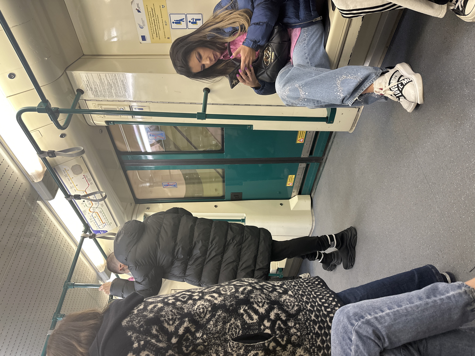 The image shows the interior of a public transport vehicle, likely a subway car, with passengers in Sofia, Bulgaria. A woman with long dark hair is sitting and looking at her mobile phone. She is wearing a dark blue puffer jacket, pink top, and light blue jeans with embellishments. On her lap rests a black quilted handbag. Her white platform sneakers are visible. Across the aisle, a person in a long black puffer coat is partially seen, leaning over, possibly interacting with a device. Another person is seated in the foreground, wearing jeans and a black and white patterned coat. The interior of the subway car is visible, including handrails, maps, and warning labels written in Bulgarian on the doors and walls.