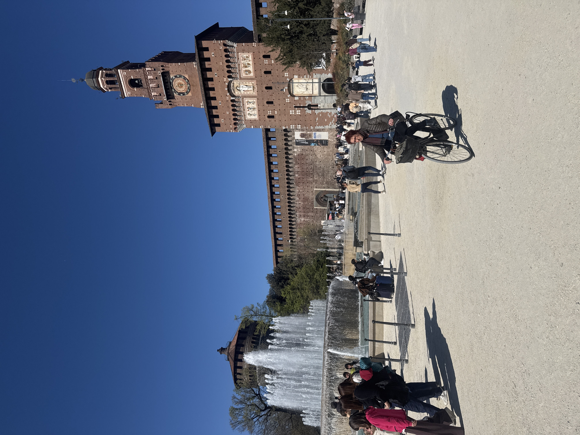 The image shows the Sforza Castle in Milan, Italy on a clear day. The castle, with its distinct architecture including a tall tower, extends into a deep blue sky. A large fountain is positioned in the foreground, with water cascading downwards. People are situated around the fountain and along the walkway, some standing, sitting, or walking. One person is riding a bicycle on the paved area adjacent to the castle. The environment includes trees and architectural details of the castle walls.