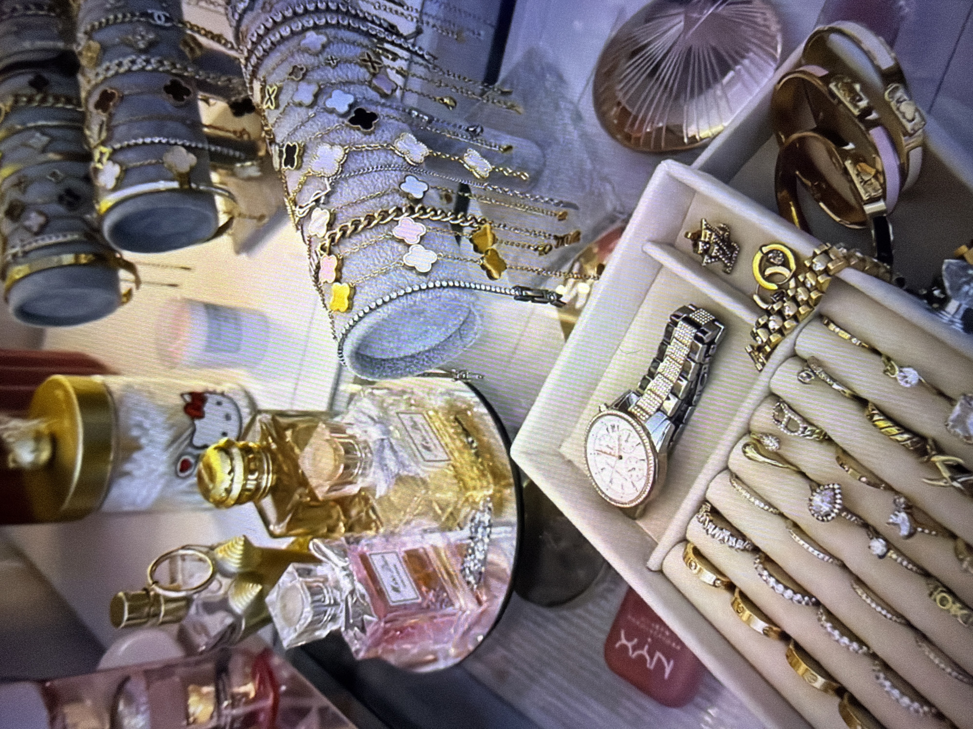 The image depicts a close-up, angled view of a vanity or display of luxury accessories, likely in Beșalma, Moldova. The scene is organized, showcasing various items arranged on flat surfaces and tiered displays.
In the foreground, a white jewelry organizer is filled with rings and a diamond-encrusted watch. The rings vary in style, from delicate bands to more elaborate pieces with prominent gemstones. The watch has a silver-toned metal band and a white face with intricate detailing. To the left of the organizer, a pink rectangular object with the "NYX" logo is partially visible.
Behind the jewelry organizer, a mirrored tray holds two perfume bottles. One bottle is pink and the other is amber-colored, both with gold caps. Beside these, a cylindrical container with a gold lid features a Hello Kitty design. Further back, multiple tiered displays hold numerous bracelets. These bracelets are adorned with charms, including clover-shaped motifs in various colors (white, black, pink, yellow) and geometric designs. Some bracelets are delicate chains, while others appear more substantial.
Other objects visible include what appears to be another perfume bottle with a gold stopper and ring, and a circular, fan-like object with a pinkish hue. The lighting in the scene is soft, highlighting the metallic and reflective surfaces of the jewelry and accessories. The overall impression is one of an organized collection of personal adornments.