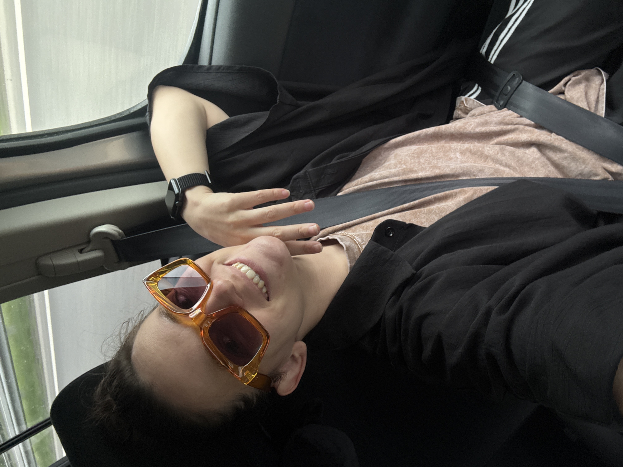 A person wearing orange sunglasses and a black watch is shown seated in a car, likely in transit. The person is wearing a black jacket and a light brown shirt underneath. A seatbelt is fastened across their body. The background shows a blurred view through the car window, suggesting motion. The location, Shali, Russia is not verifiable from this image.