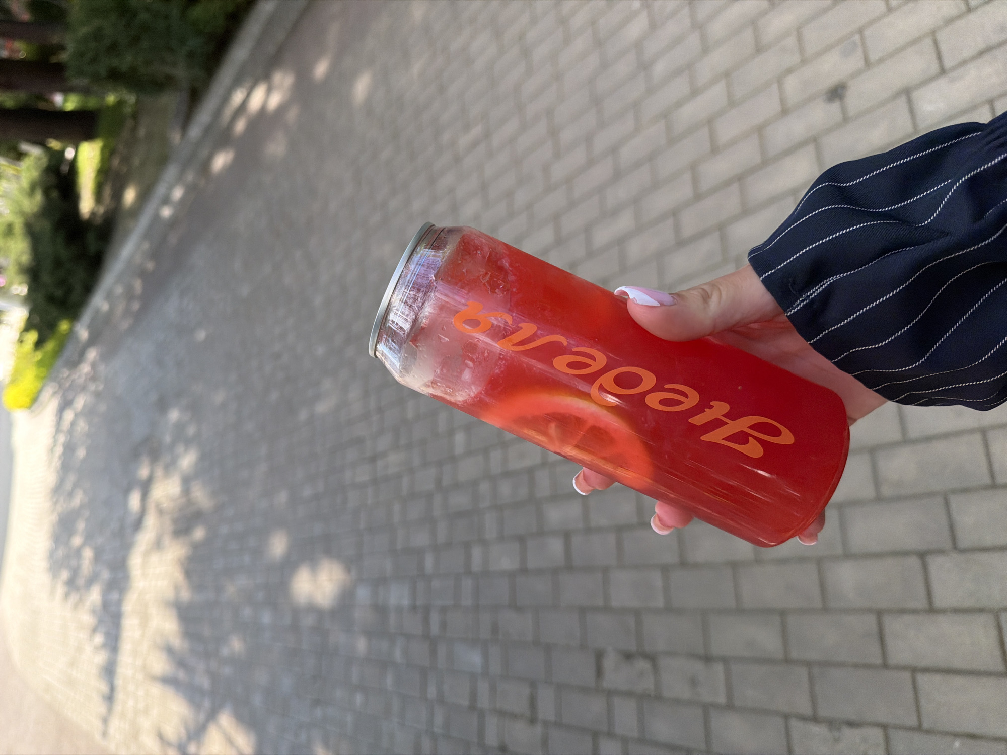 A person is holding a clear glass-like beverage container filled with a red-colored liquid and a slice of fruit, likely lemon, with an orange logo displayed. The container's top has a metallic silver-colored rim. The person holding the container has fingernails painted with a white and pink design and is wearing a dark-colored shirt with white stripes. The background consists of a brick-paved surface and blurry trees. The exact location in Kazanlak, Bulgaria, is not discernible from the image alone.
