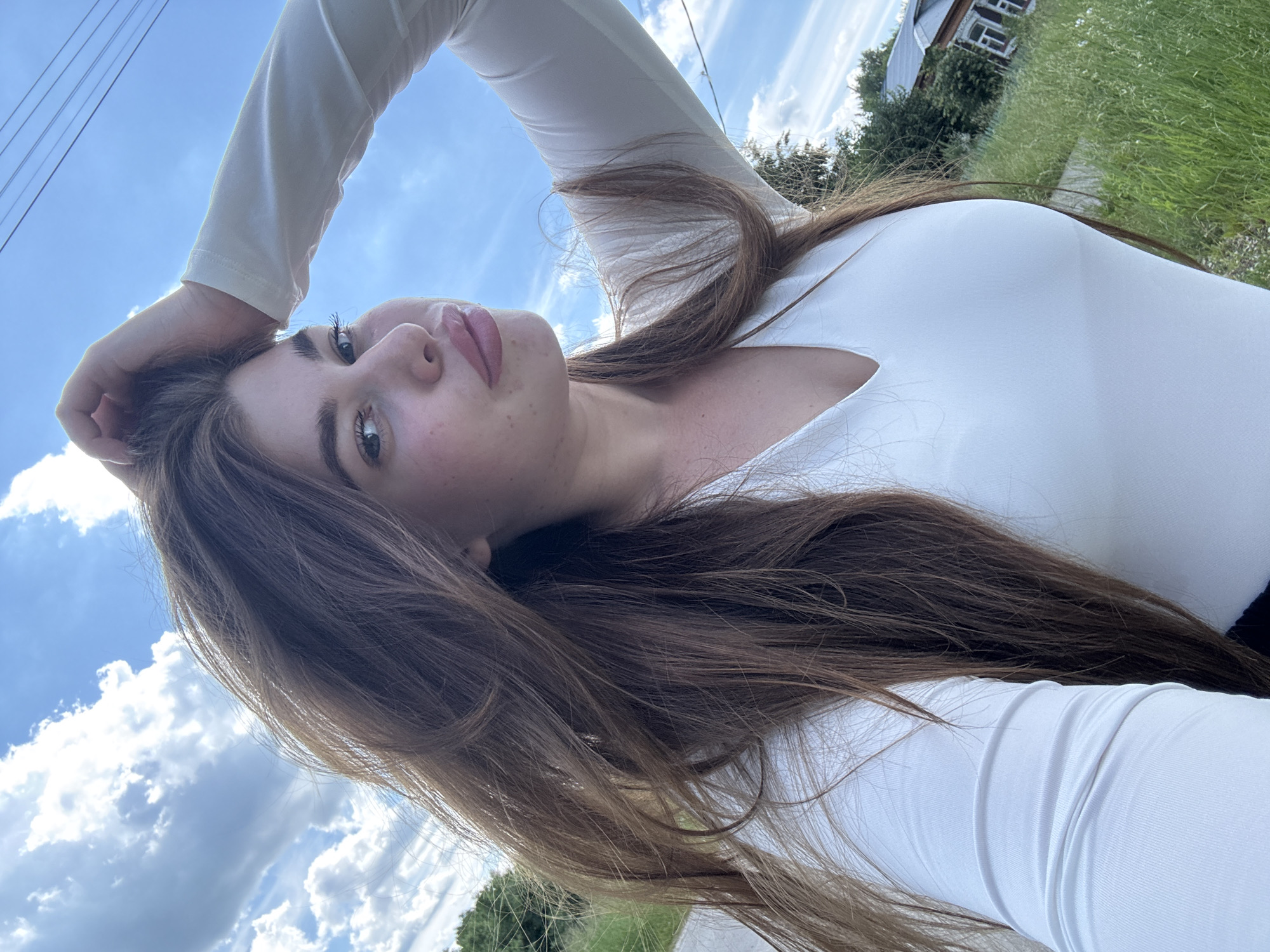A young woman with long, light brown hair and fair skin is pictured from the chest up. She is wearing a white, long-sleeved top and has her right arm raised, with her hand touching her head. Her gaze is directed towards the camera, and she has plump lips. The background shows a bright blue sky with white clouds, power lines overhead, and a blurred green landscape with a rustic house visible on the right side. This scene is suggestive of an outdoor location, potentially in a rural area or the outskirts of a town in Мещёры, Russia, given the presence of the traditional wooden house and the general natural environment.