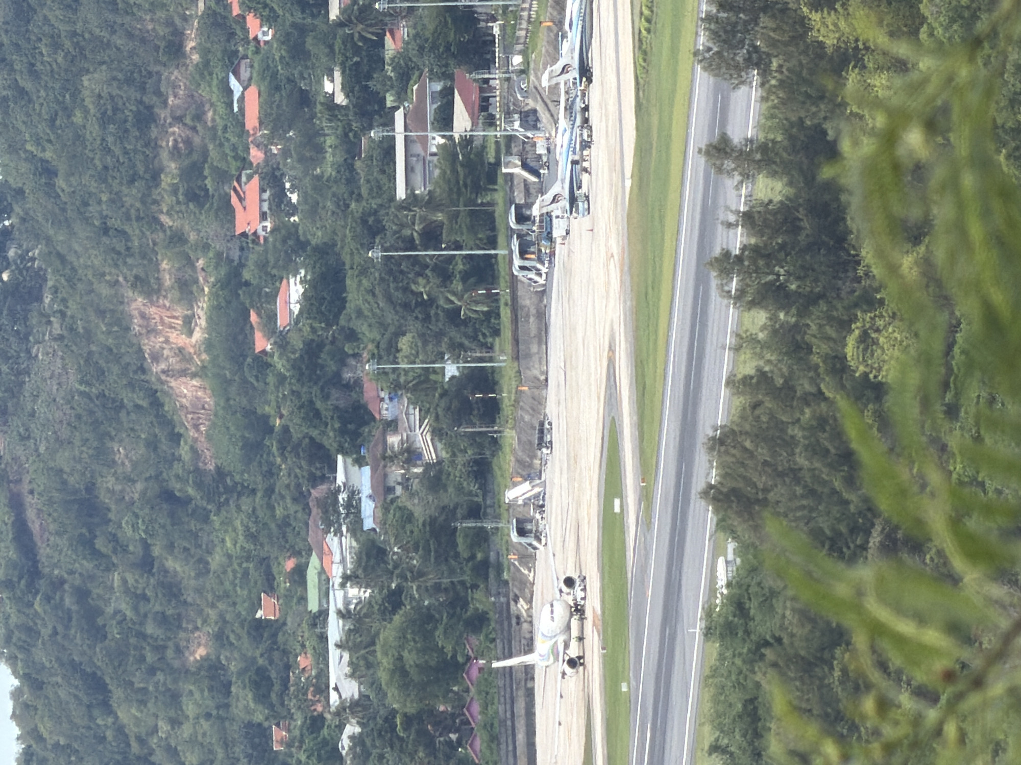 An aerial view captures an airport tarmac in Ban Bang Rak, Thailand, with a lush, hilly landscape and scattered residential buildings in the background. In the foreground, a white airplane with colorful markings is parked on the tarmac, surrounded by small vehicles and a group of people. Further down the tarmac, several other airplanes are visible, some of which appear to be connected to transport vehicles. The airport infrastructure, including light poles and stairs, is spread across the concrete surface. The surrounding area is densely covered with trees and vegetation, with glimpses of red-tiled roofs of houses peeking through the foliage. A paved runway is in the lower part of the image, separated from the tarmac by a grassy area.