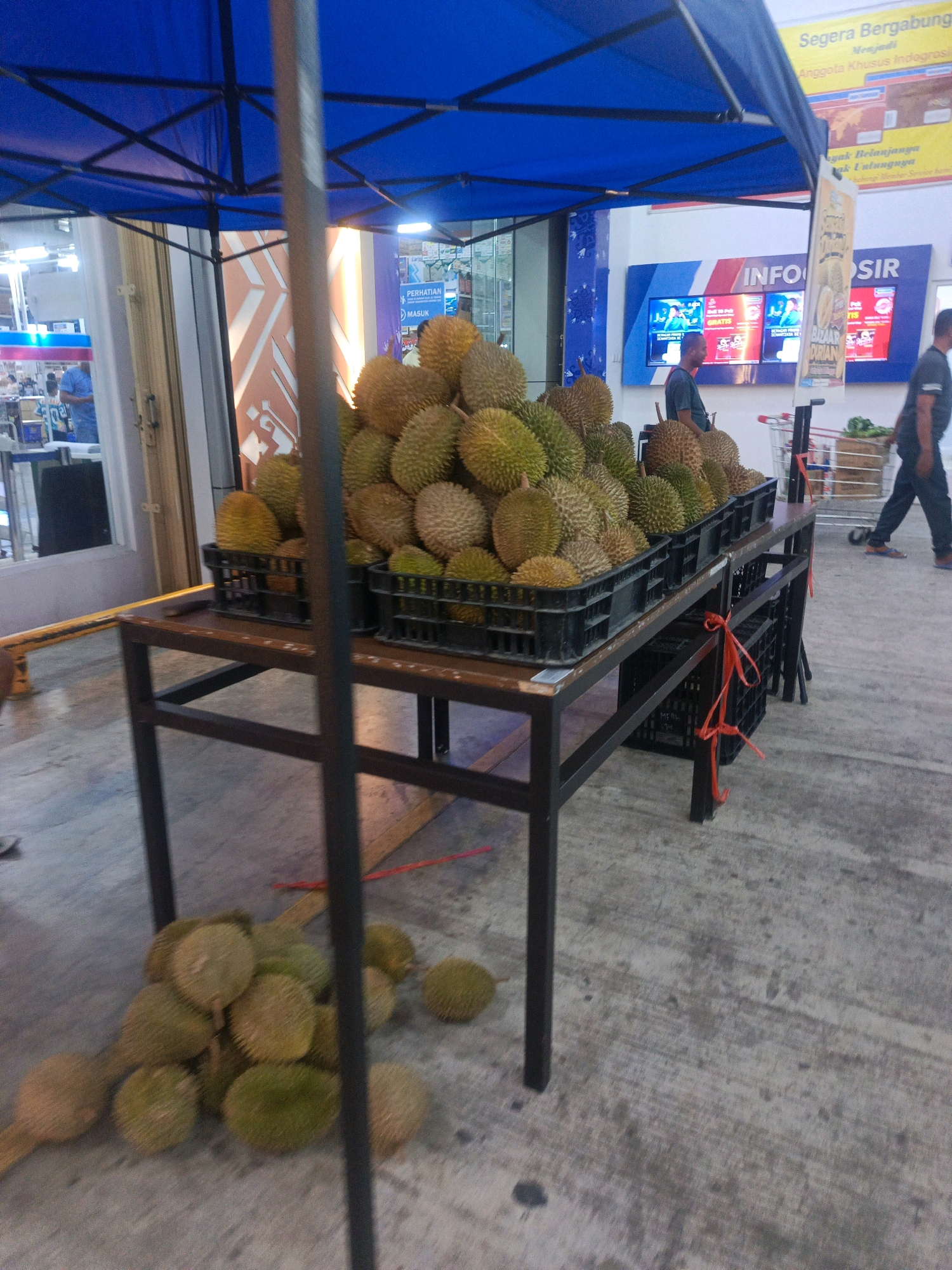 In the city of Gorontalo, Indonesia, a stall is set up under a blue canopy. The stall displays a large quantity of durian fruit, some of which are placed in black plastic crates on a dark metal table. Additional durian fruits are piled on the ground in front of the table. The background shows a building facade with various advertisements and illuminated screens. Two men are visible; one is standing near the durian display, and another is walking away from the camera towards the right side of the image. A shopping cart with a basket containing produce is positioned behind the durian display. The ground is a concrete surface.