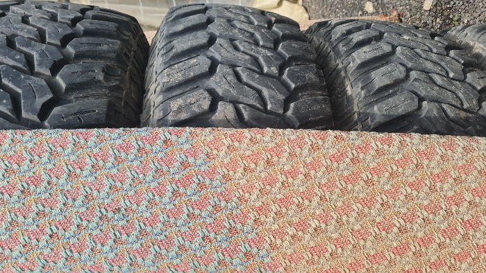 In Mate Asher Regional Council, Israel, the image presents a static, vertically oriented scene divided into two primary visual elements. The left half is occupied by a textured, vertical surface displaying a repetitive geometric pattern composed of small, interwoven shapes in pink, light blue, and beige. The right half features a vertical stack of at least four large, dark grey off-road tires. These tires exhibit aggressive, blocky tread patterns and appear weathered and covered in dust and dirt. A partial, indistinct background behind the tires suggests an outdoor, possibly industrial or storage environment, with hints of a lighter-colored structure and ground debris. No individuals or active interactions are depicted.