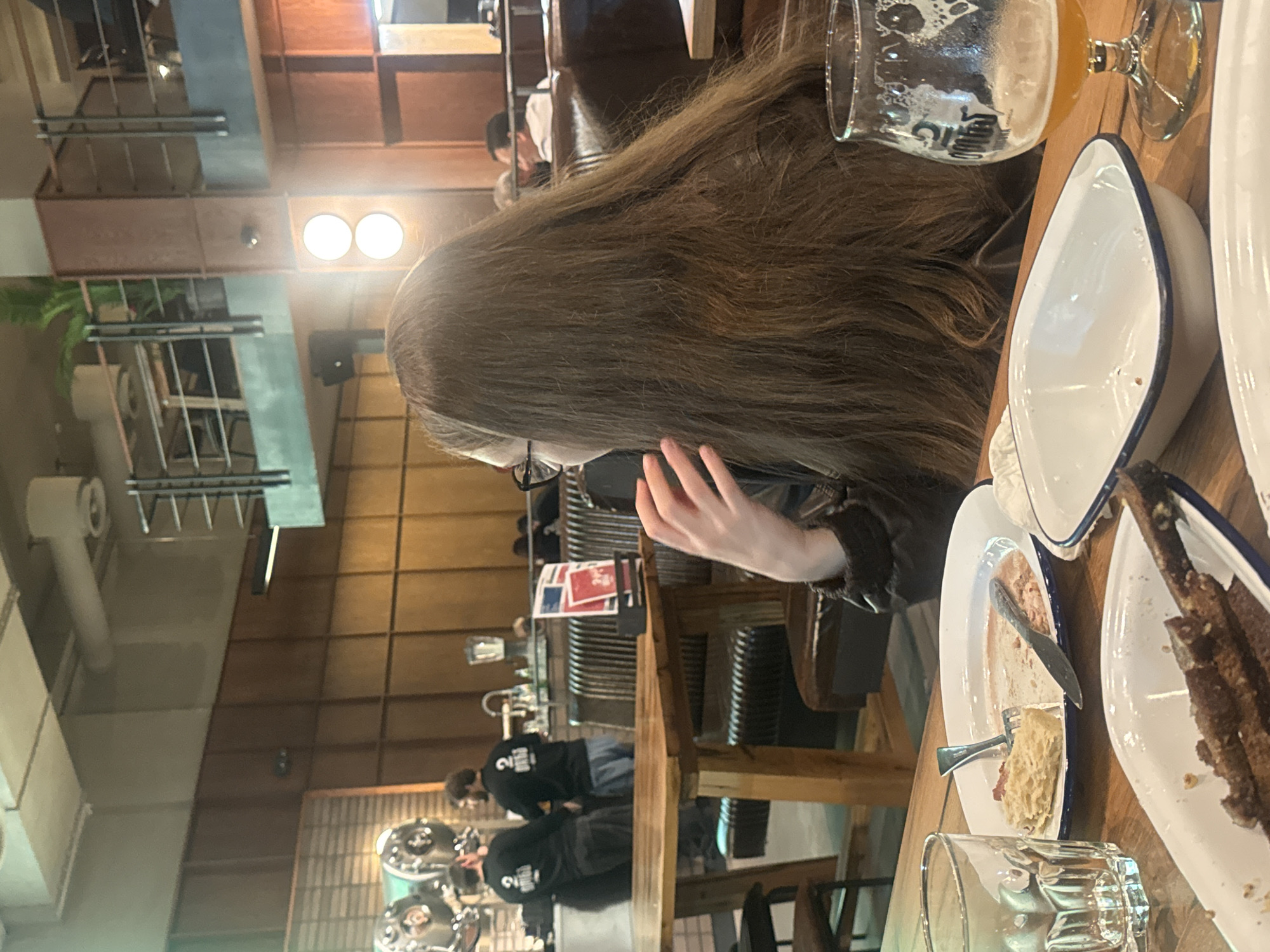 A long-haired woman wearing glasses is seated at a wooden table in a restaurant. She is holding a cell phone to her ear. The table is set with empty white plates with blue rims, cutlery, and partially consumed beverages. In the background, restaurant staff are visible and the interior features wooden walls and hanging light fixtures. The scene is set in a restaurant in Tbilisi, Georgia.
